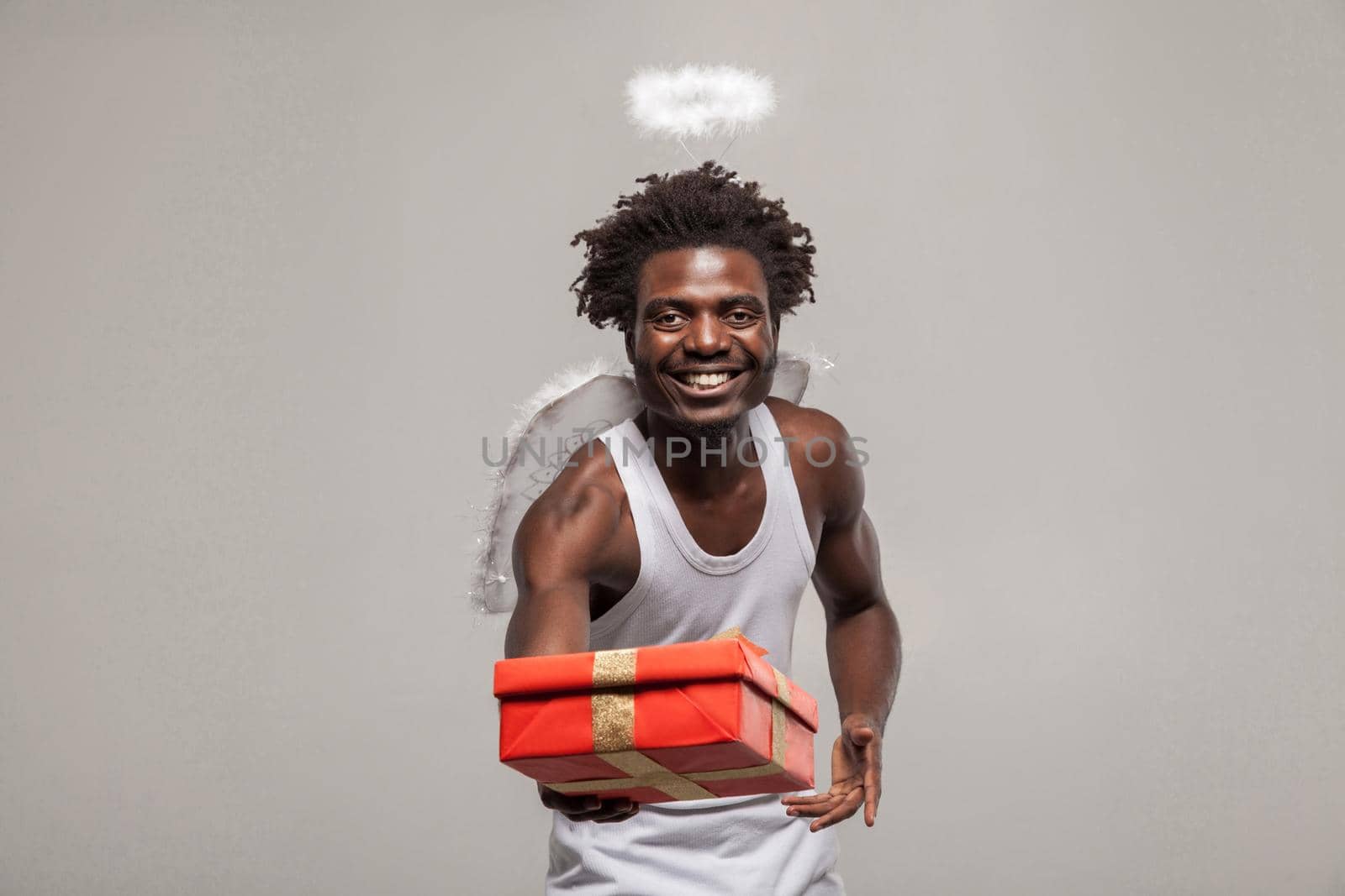 Love concept and sharing goft. dark skinned bearded African angel funny man with white shirt, nimbus and winks atanding and holding present gift box. its for you. Indoor, isolated on gray background