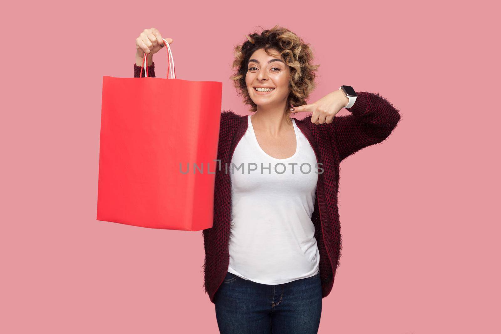 Cheerful beautiful young woman in blue shirt with curlty hairstyle standing and pointing finger to shopping bags and toothy smile, looking at camera. Studio shot, pink background, isolated, indoor