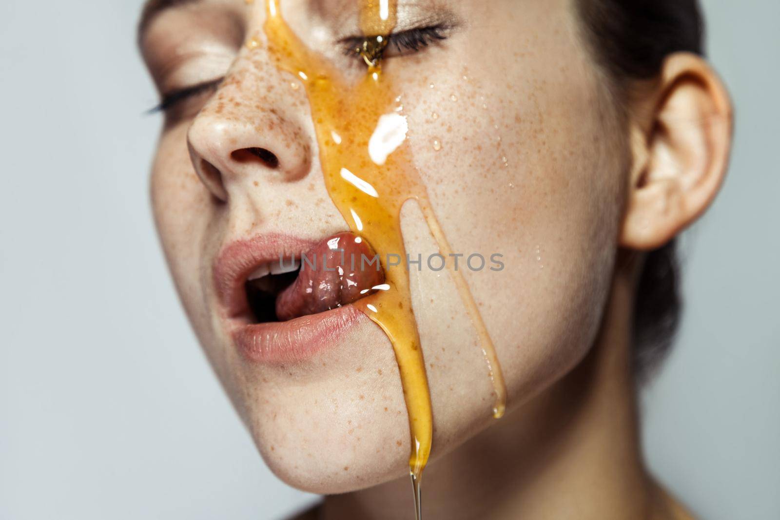 Closeup portrait of beautiful young brunette woman with freckles and honey on face, tongue out and licking with closed eyes and passion. indoor studio shot isolated on gray background.