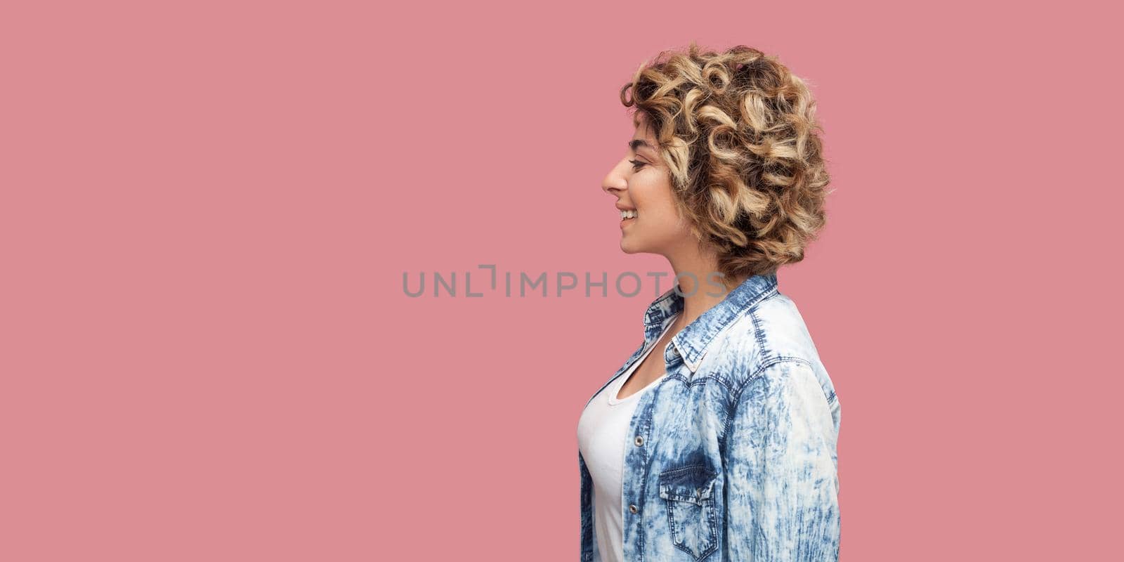 Profile side view portrait of young happy woman with makeup and blonde curly hairstyle in casual blue shirt standing, looking forward with toothy smile. indoor studio shot, isolated on pink background