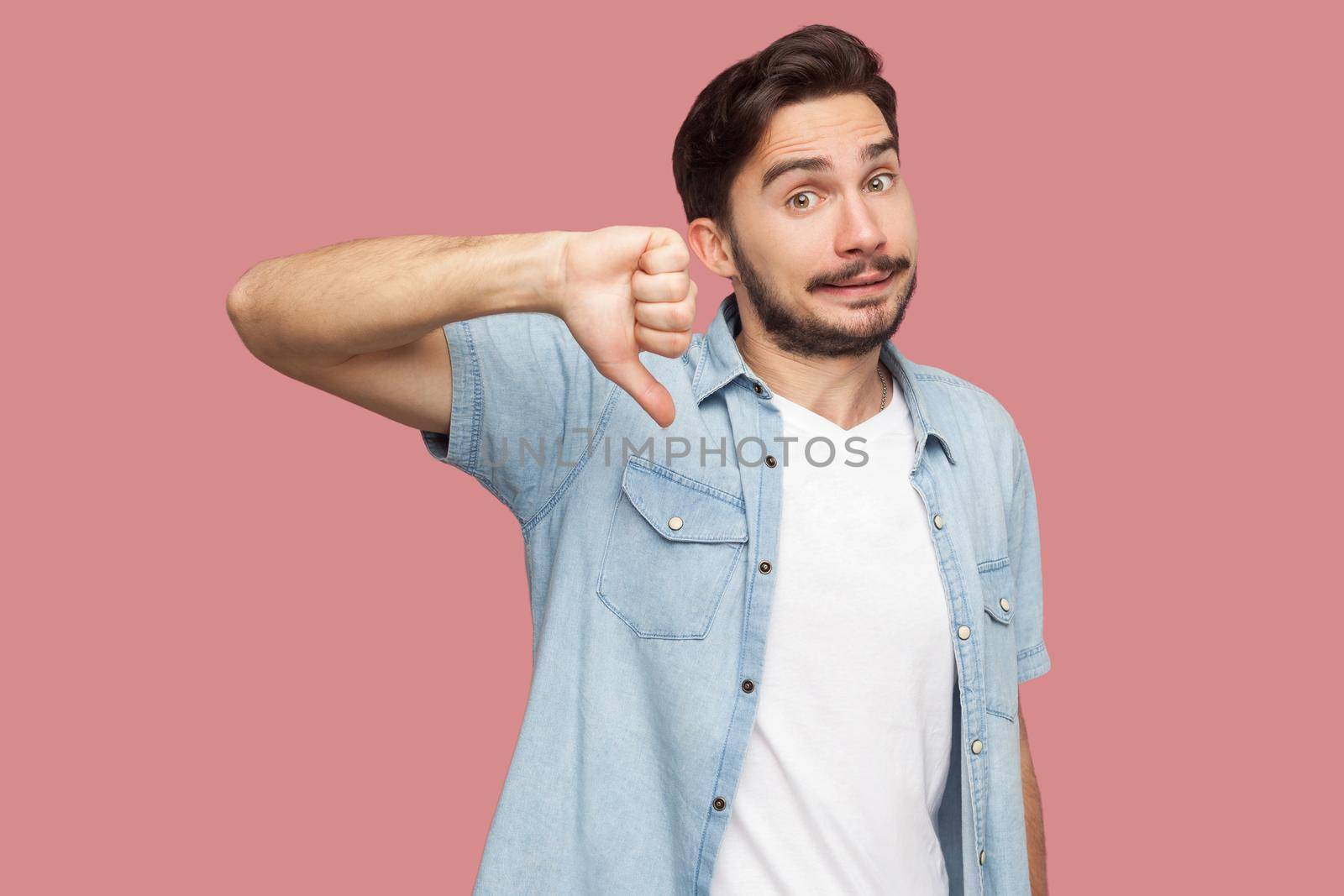 No, Dislike. Portrait of dissatisfied handsome bearded young man in blue casual style shirt standing, thumbs down and looking at camera . indoor studio shot, isolated on pink background.