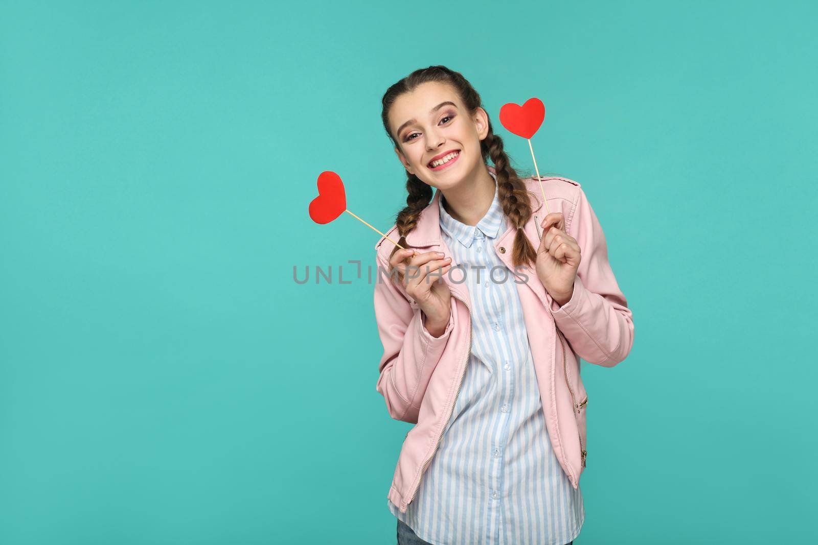 Happy beautiful girl in casual style, pigtail hairstyle and pink jacket, standing and holding red heart stickers and looking at camera and toothy smiling, Indoor, isolated on blue or green background