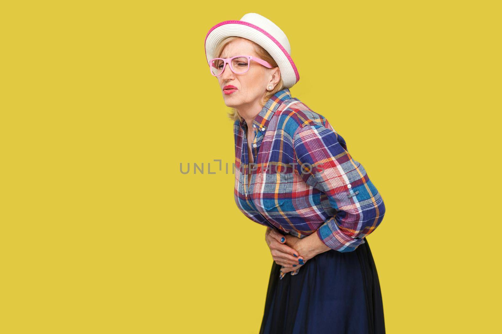 Stomach pain. Profile side view portrait of modern stylish mature woman in casual style with hat and eyeglasses standing and holding her painful belly. indoor studio shot isolated on yellow background