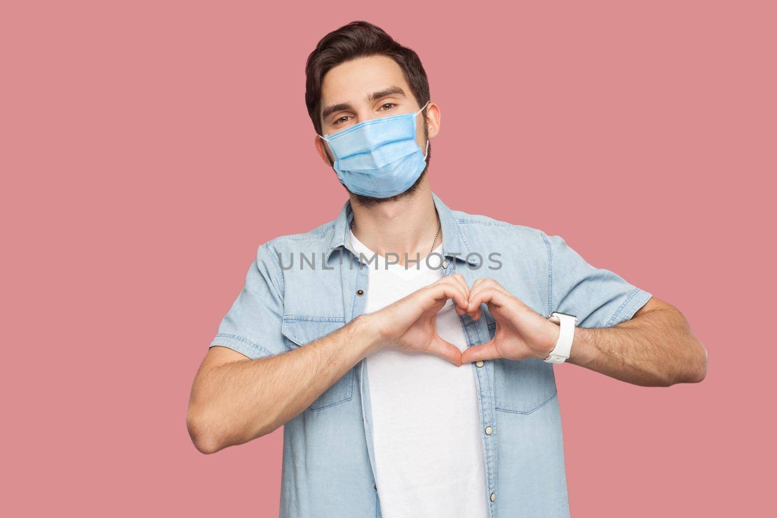Portrait of happy smiley young man with surgical medical mask in blue shirt standing with love hand gesture on heart and looking at camera smiling. indoor studio shot, isolated on pink background.