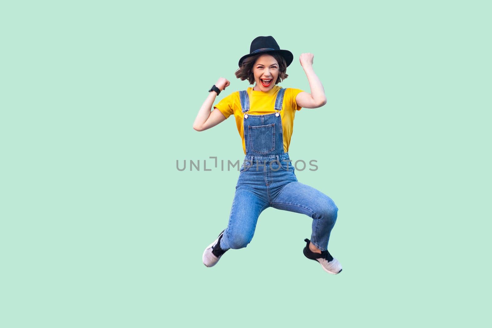 Portrait of screaming happy suprised young girl in blue denim overalls, yellow shirt and black hat jumping, looking and celebraiting her victory. indoor studio shot isolated on light green background.