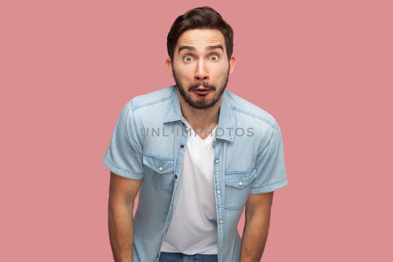 Portrait of surprised handsome bearded young man in blue casual style shirt standing and looking at camera with shocked unbelievable face. indoor studio shot, isolated on pink background.