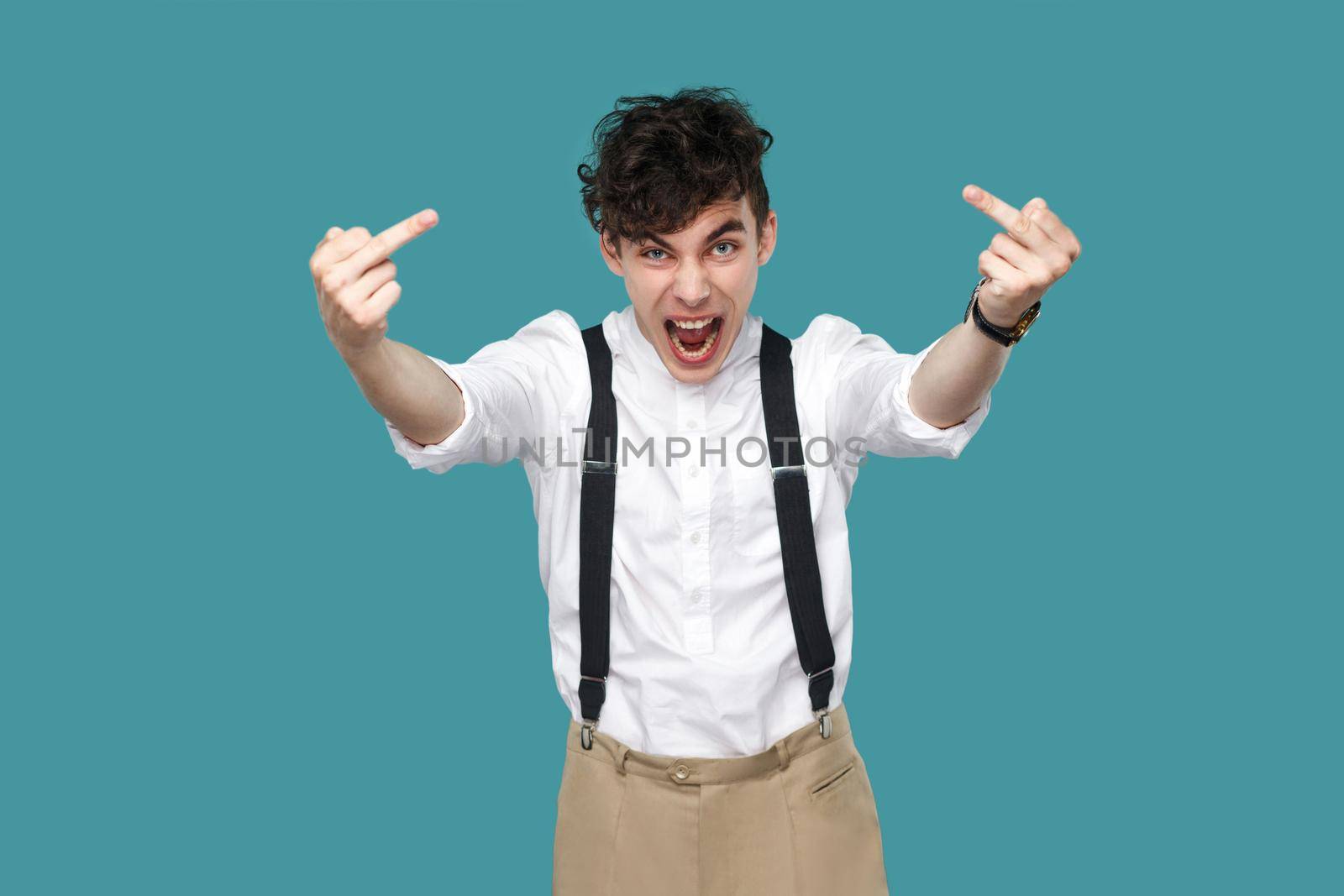 Angry man screaming and showing middle finger fuck sign. Portrait of handsome hipster curly young businessman in classic casual white shirt, suspender. indoor studio shot isolated on blue background