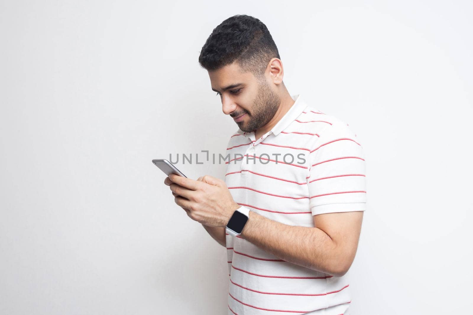 Side view portrait of happy handsome blogger young adult man wearing T-shirt standing, using smartphone and texting message with smile. Indoor, isolated, studio shot, white background, copy space