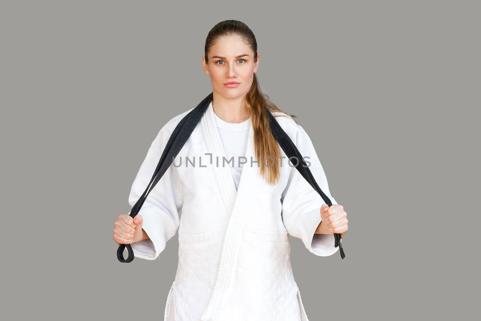 Confident attractive athletic woman in white kimono standing and wraps black belt around the neck, looking at camera. Japanese karate and sports concept. Indoor, studio shot, isolated, grey background