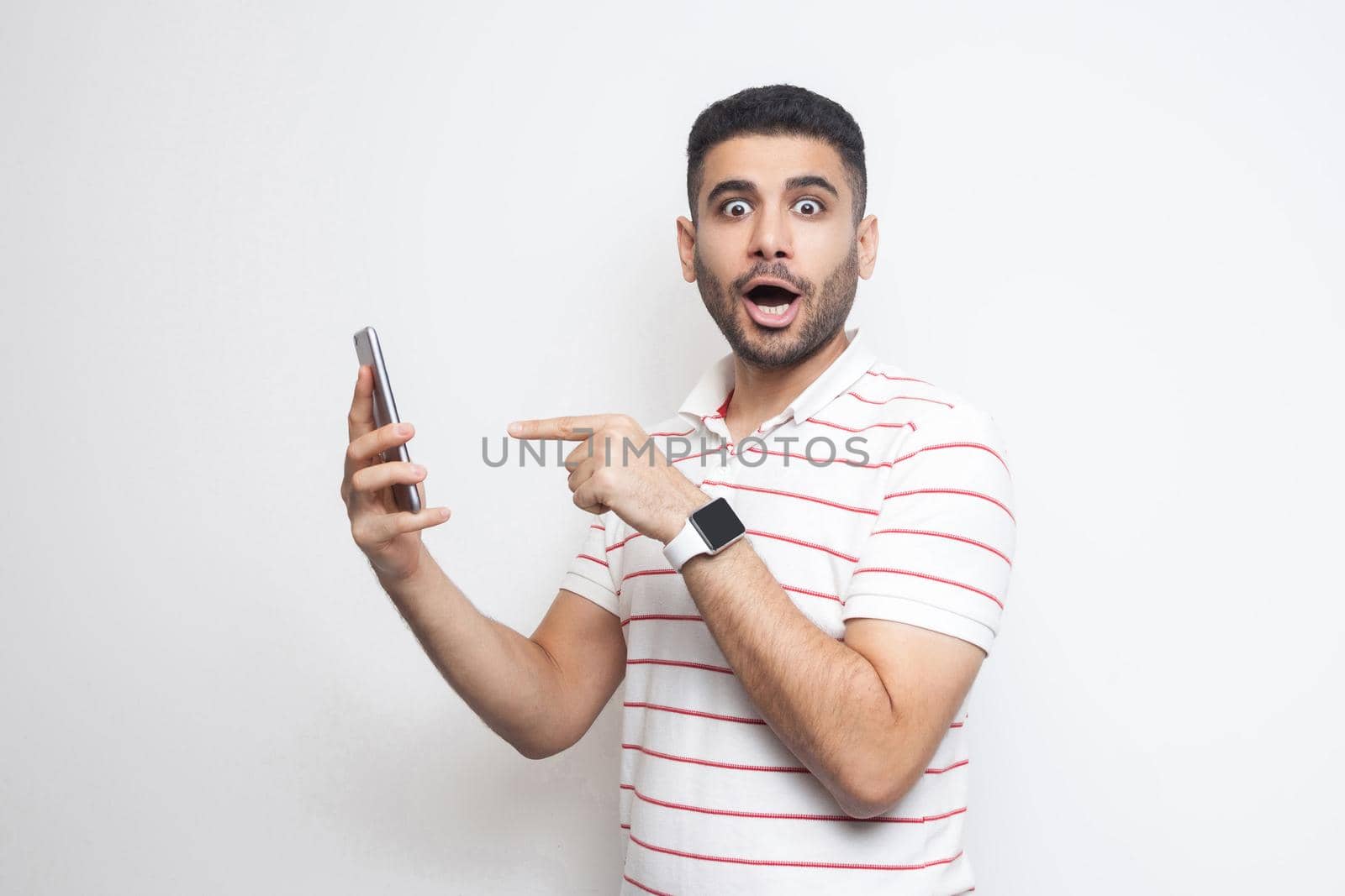 Portrait of shocked young adult man in white t-shirt standing, holding phone and pointing finger with suprised face and big eyes. Indoor, isolated, studio shot, white background, looking at camera