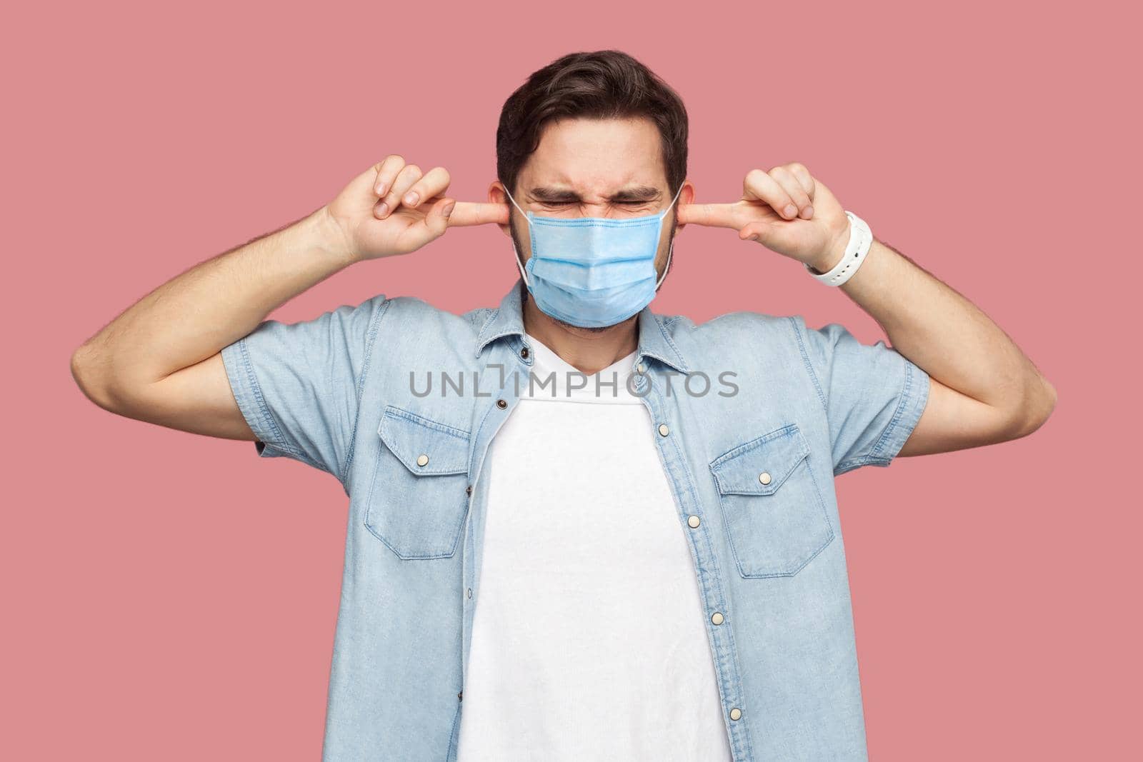 I don't want to hear you. Portrait of confused young man with surgical medical mask in blue shirt standing and putting fingers on ears and dont care. indoor studio shot, isolated on pink background