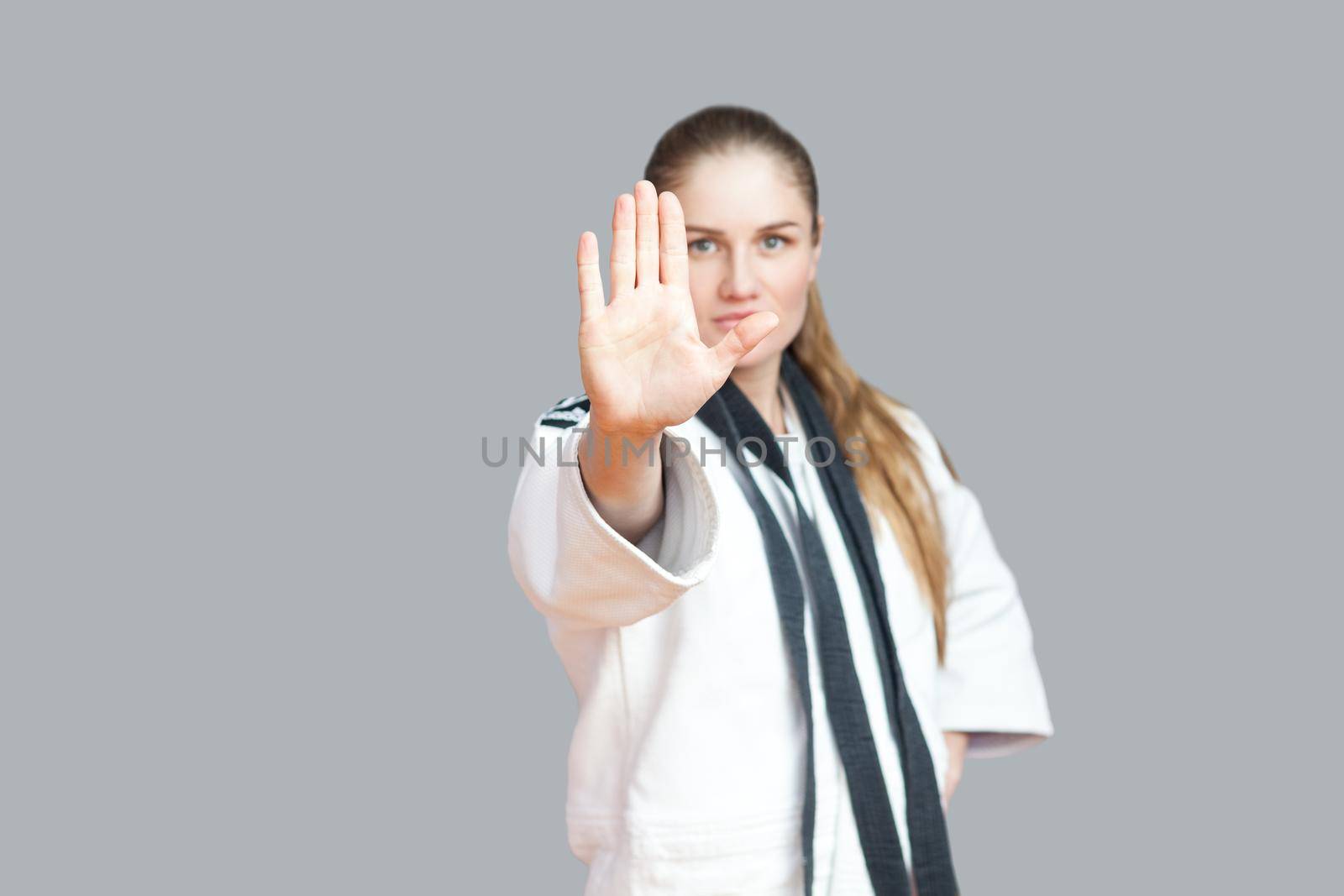 Agressive young athletic woman in white kimono with black belt wraps around the neck standing and stopping you. Japanese karate and sports concept. indoor studio shot, isolated, grey background