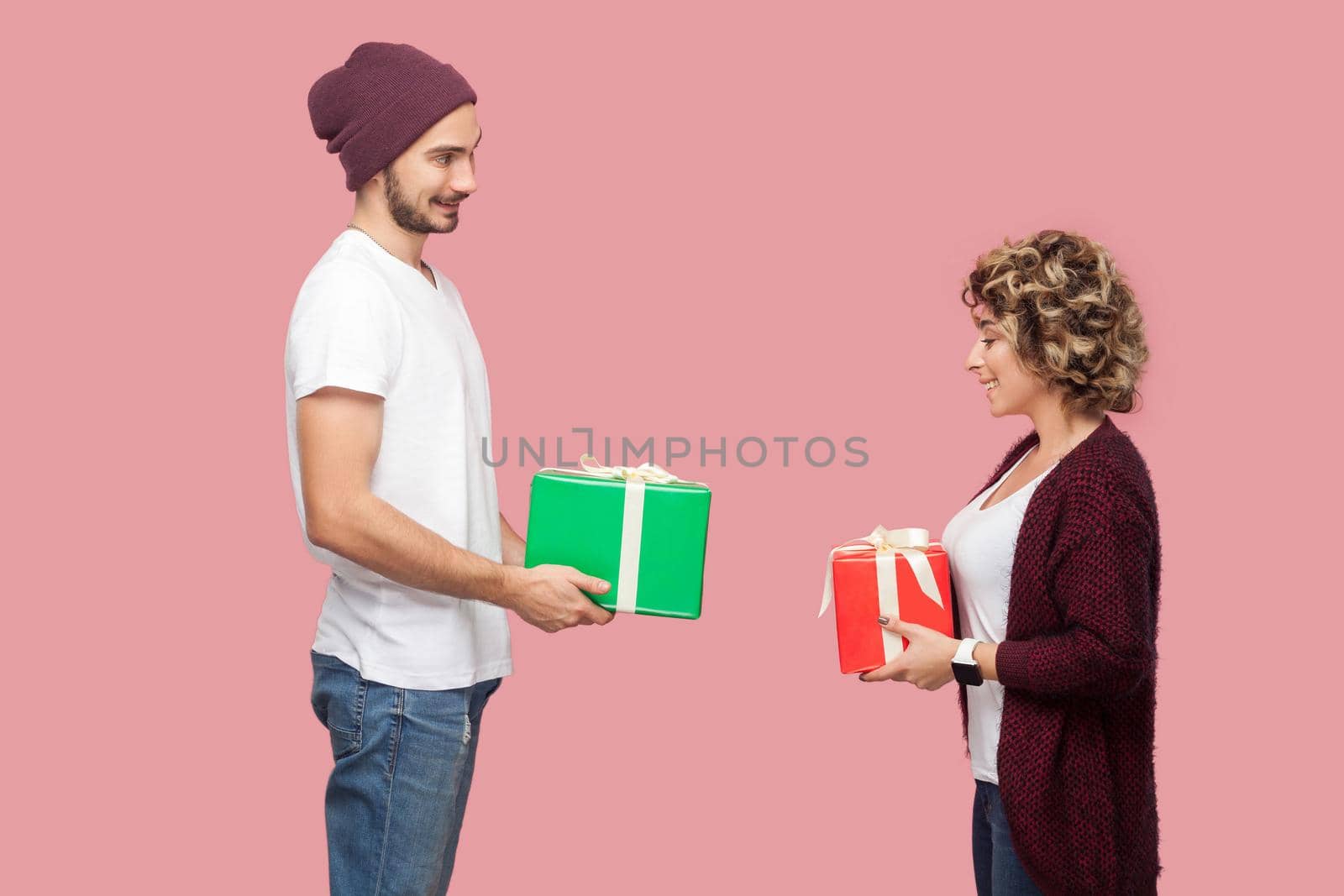 Side view portrait of suprised couple of friends in casual style standing, giving present boxes to each other, celebrate anniversary, toothy smile. Isolated, indoor, studio shot, pink background