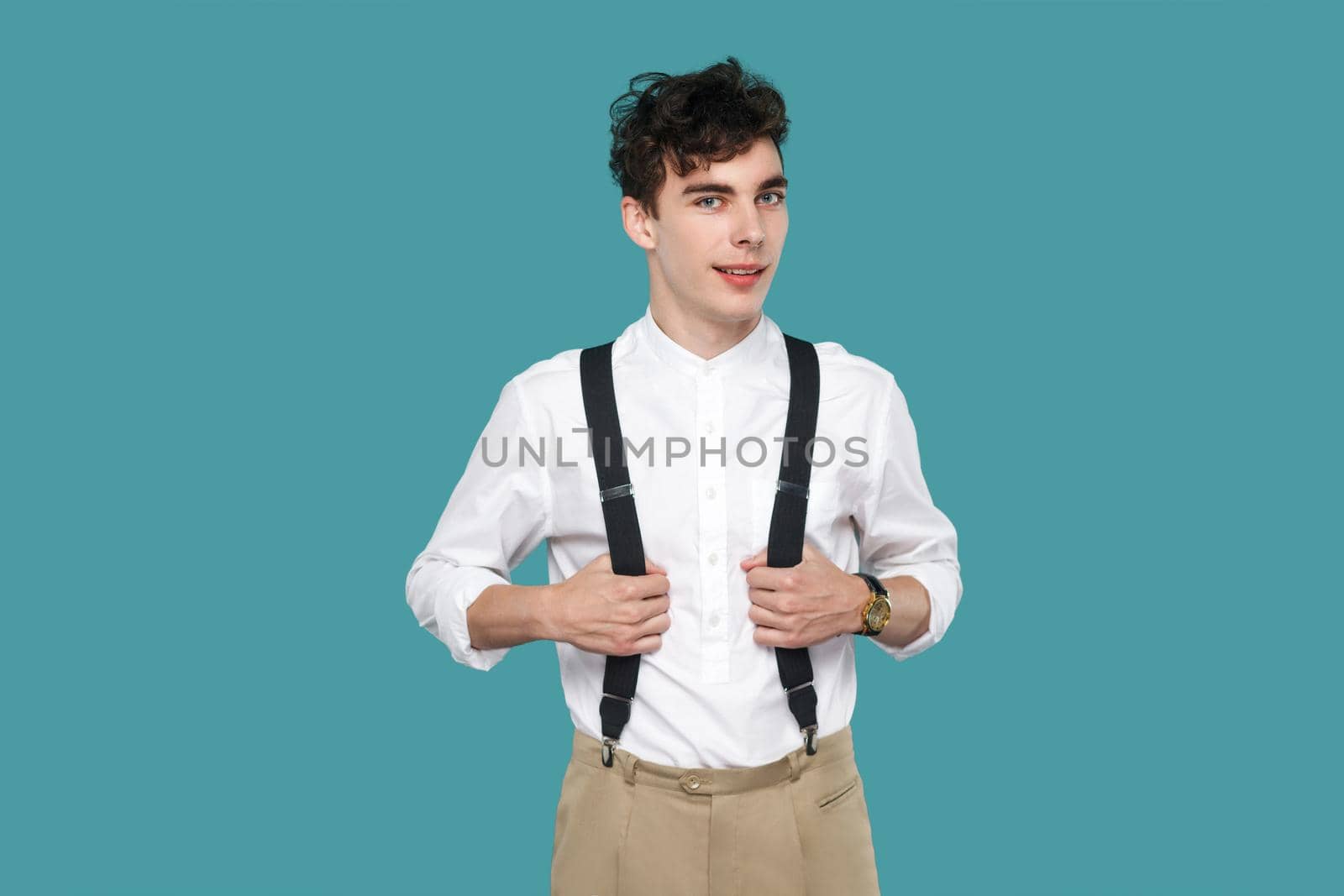 happy man toothy smile, standing and looking at camera. Portrait of handsome hipster curly young man in classic casual white shirt and suspender. indoor studio shot isolated on blue background.