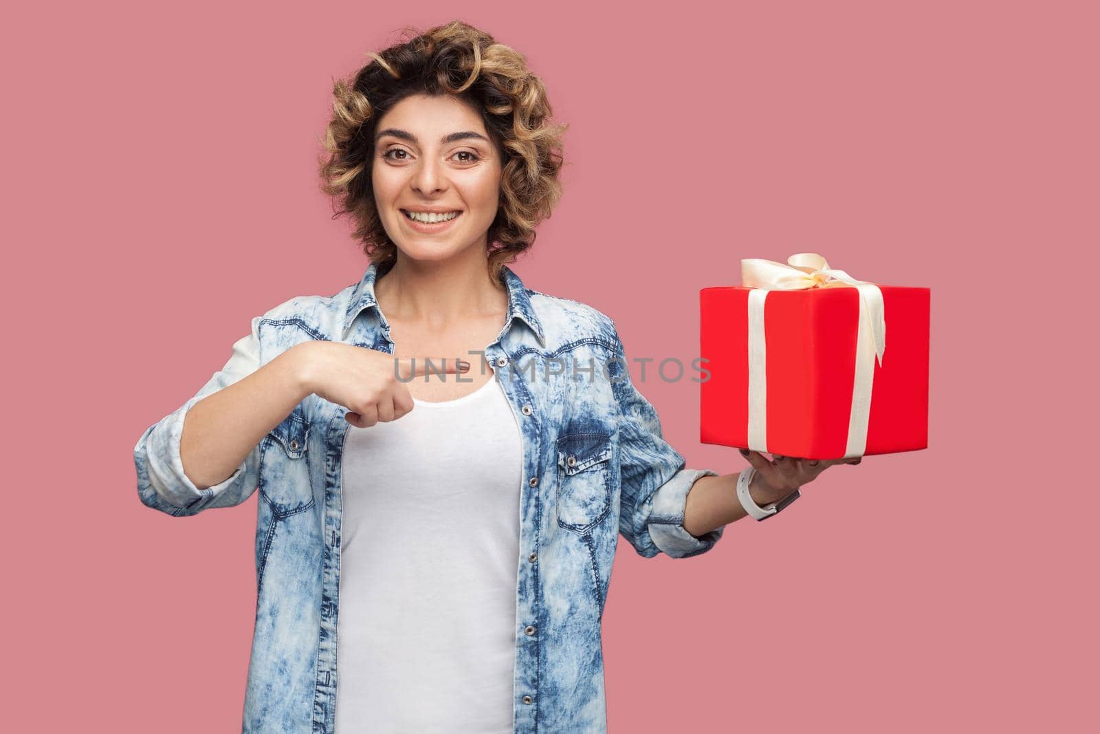 Portrait of happy young woman in blue shirt with curlty hairstyle holding red gift box and pointing finger, looking at camera. Indoor, isolated, studio shot, copy space, pink background