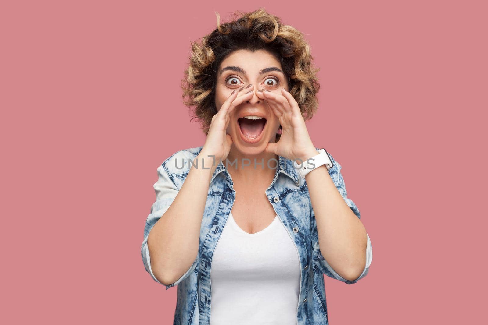 Portrait of shocked young woman with curly hairstyle in casual blue shirt standing, screaming, sharing something and looking at camera with amazed face. indoor studio shot, isolated on pink background
