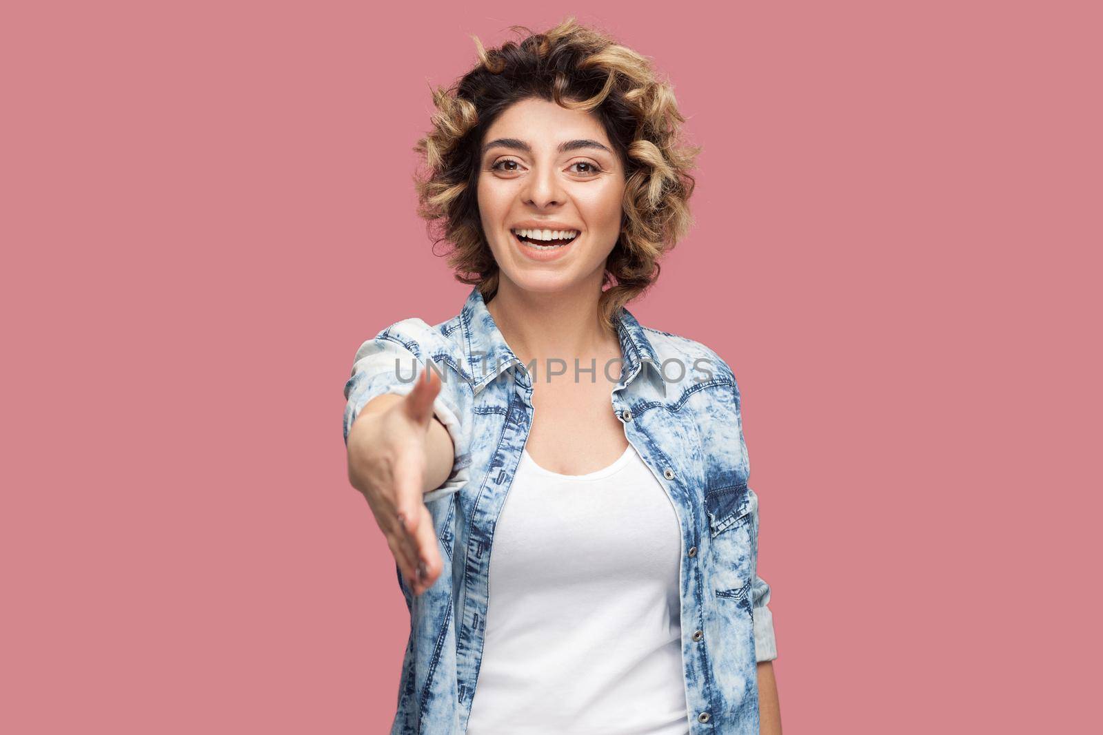 Portrait of happy young woman with curly hairstyle in casual blue shirt standing, toothy smile, looking and giving her hand to greeting and handshake. indoor studio shot, isolated on pink background.