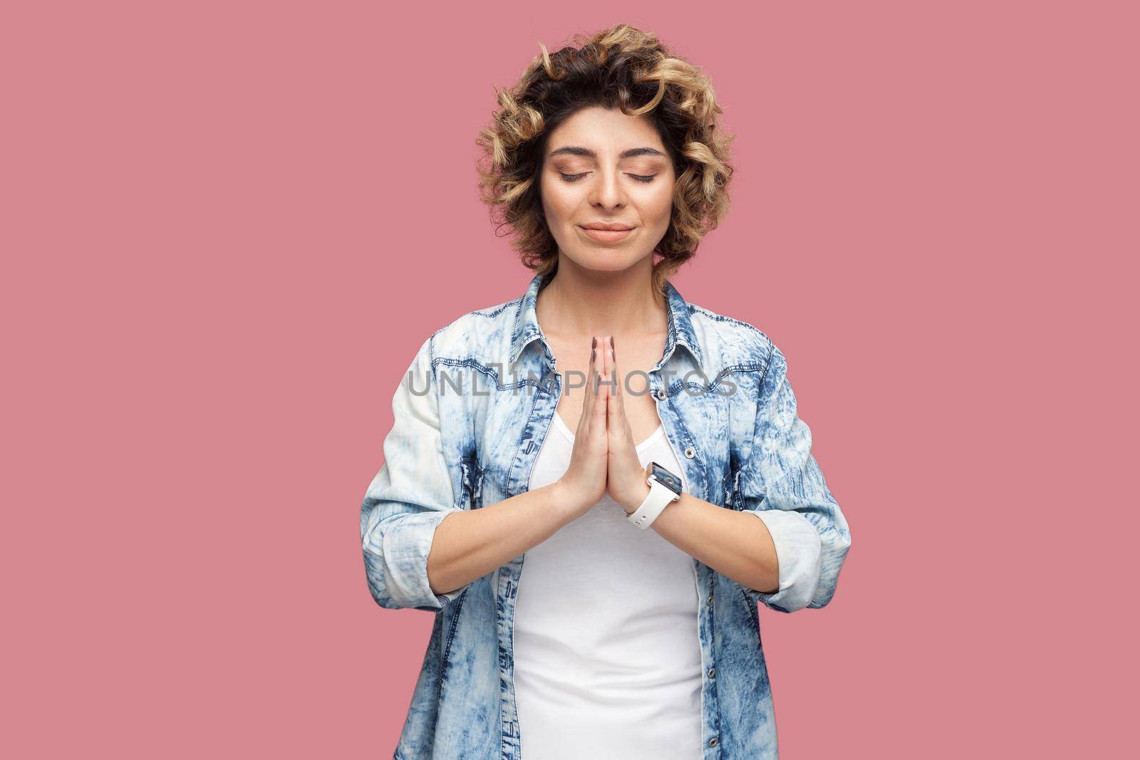 Portrait of calm young woman with curly hairstyle in casual blue shirt standing with closed eyes, palm hands, smile and doing yoga meditating. indoor studio shot, isolated on pink background.