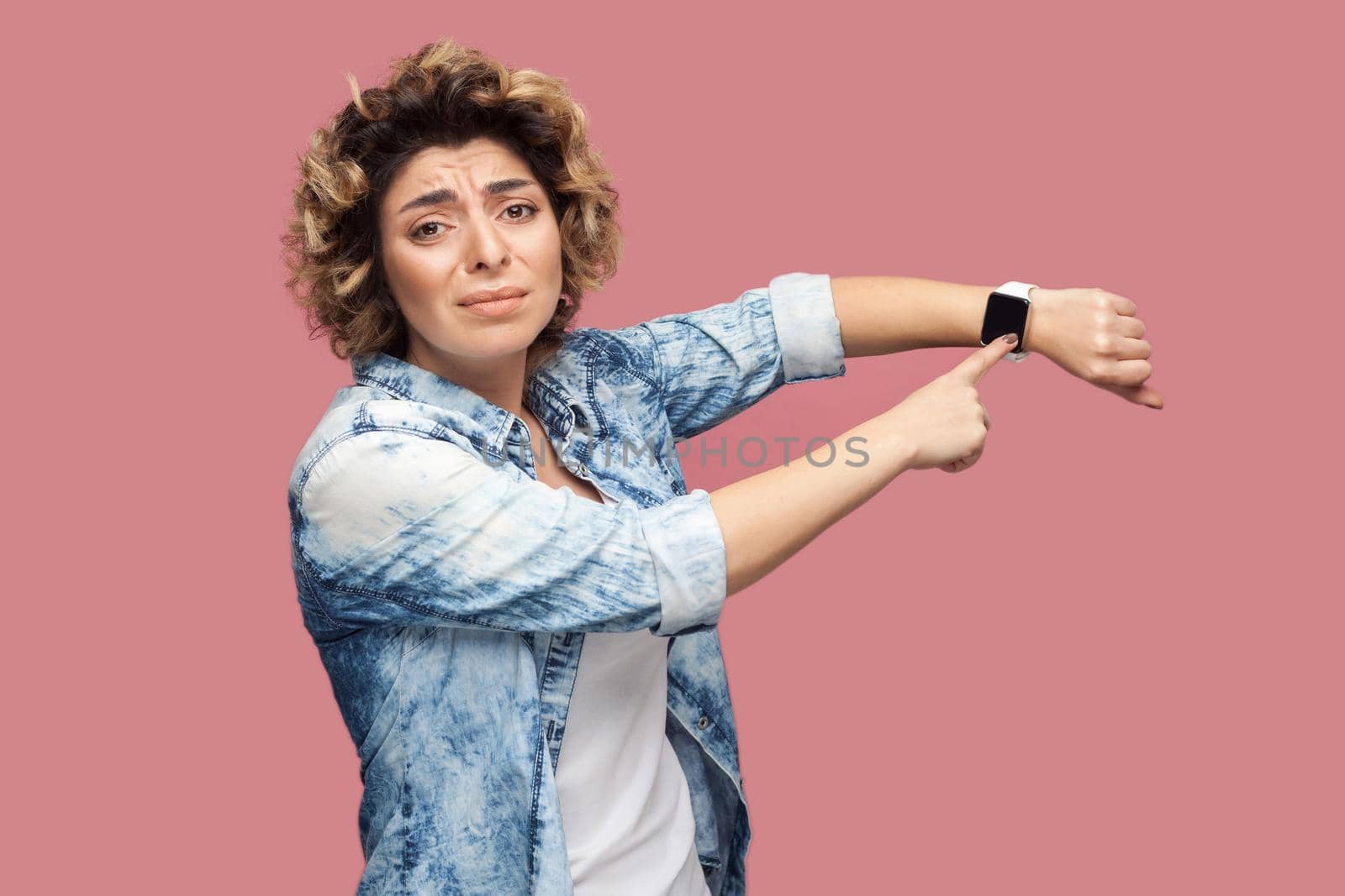I need more time. Portrait of young pleased woman with curly hairstyle in casual blue shirt standing and pointing at her smart watch and want more time. indoor studio shot, isolated on pink background