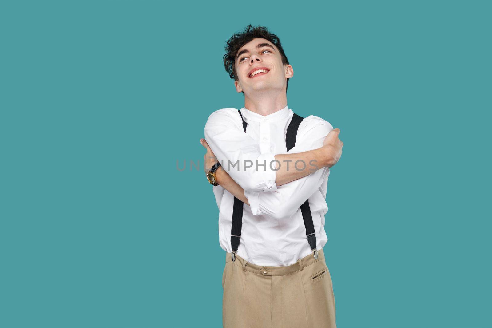 Happy excited man toothy smile and hug himself. Portrait of handsome hipster curly young businessman in classic casual white shirt, suspender standing. indoor studio shot isolated on blue background