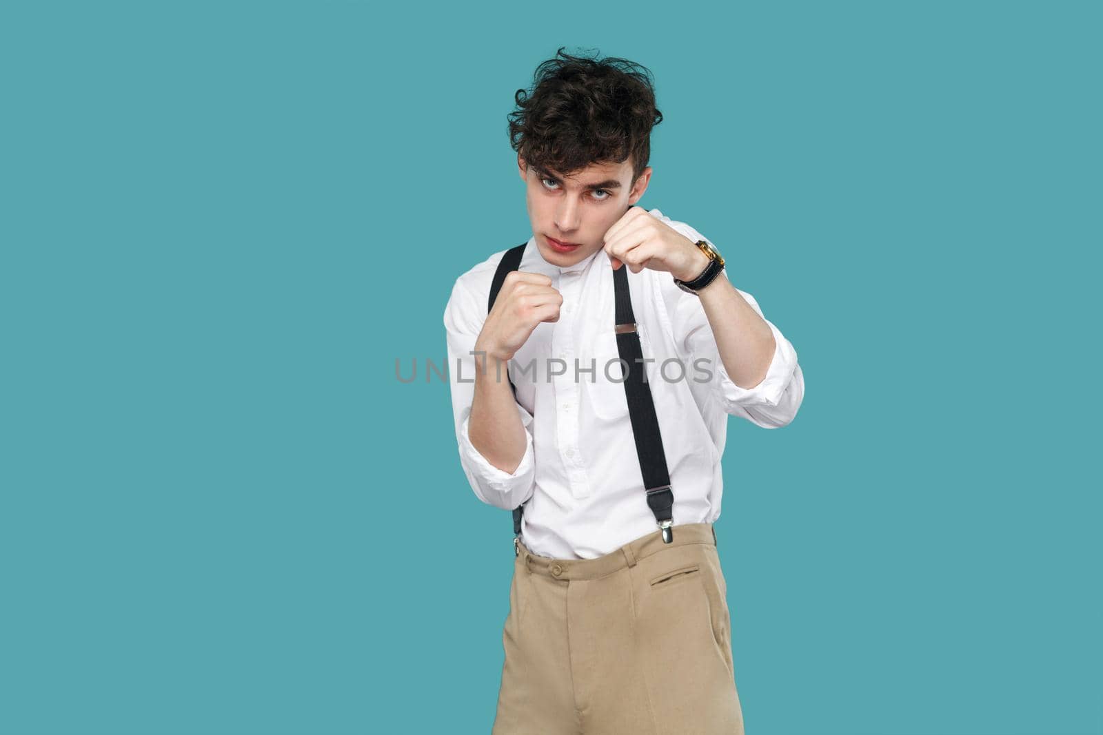 Serious man with boxing fist looking at camera. Portrait of handsome hipster curly young businessman in classic casual white shirt, suspender standing. indoor studio shot isolated on blue background.