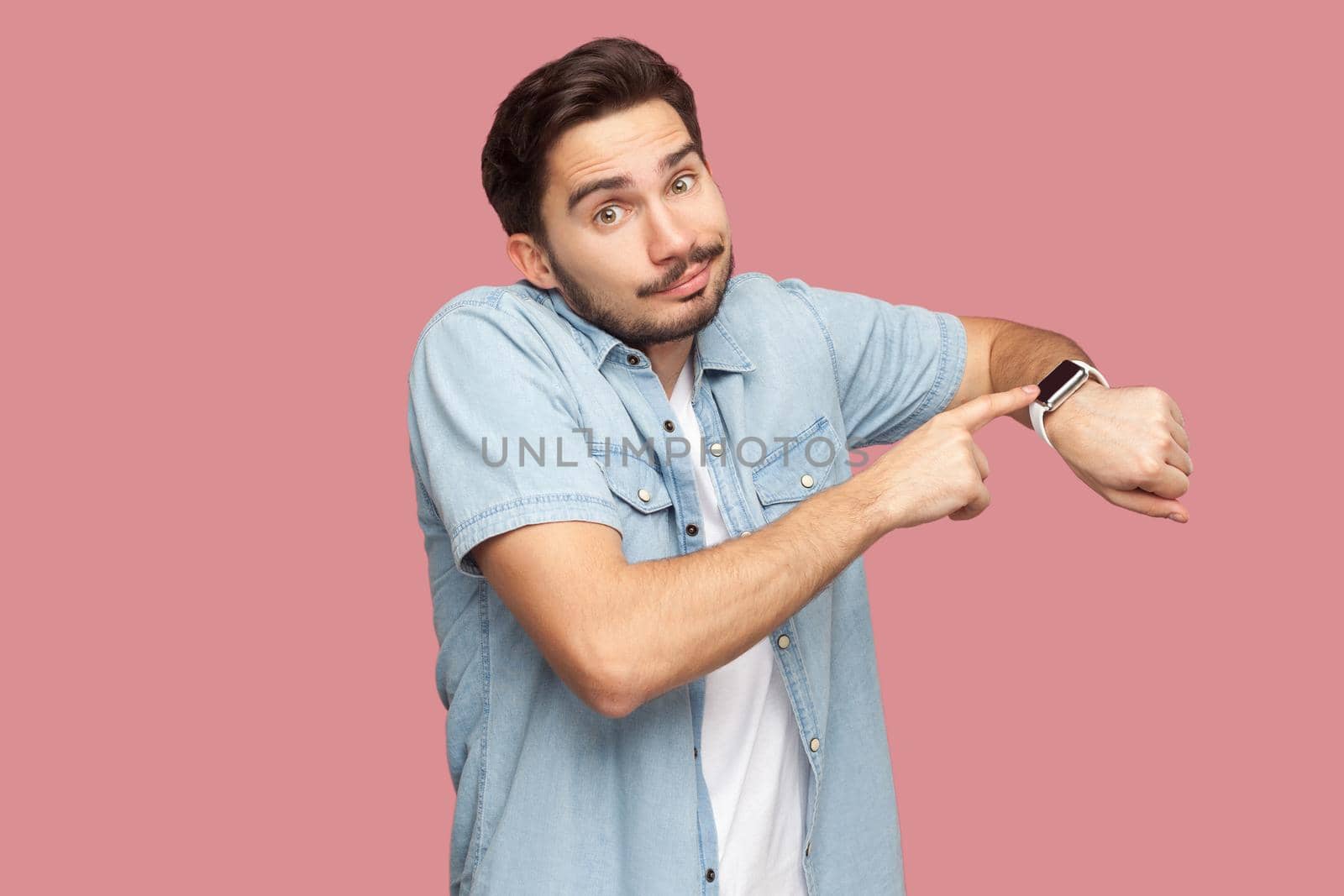 Portrait of sad handsome bearded young man in blue casual style shirt standing pointing and showing his smart watch and looking at camera. indoor studio shot, isolated on pink background.