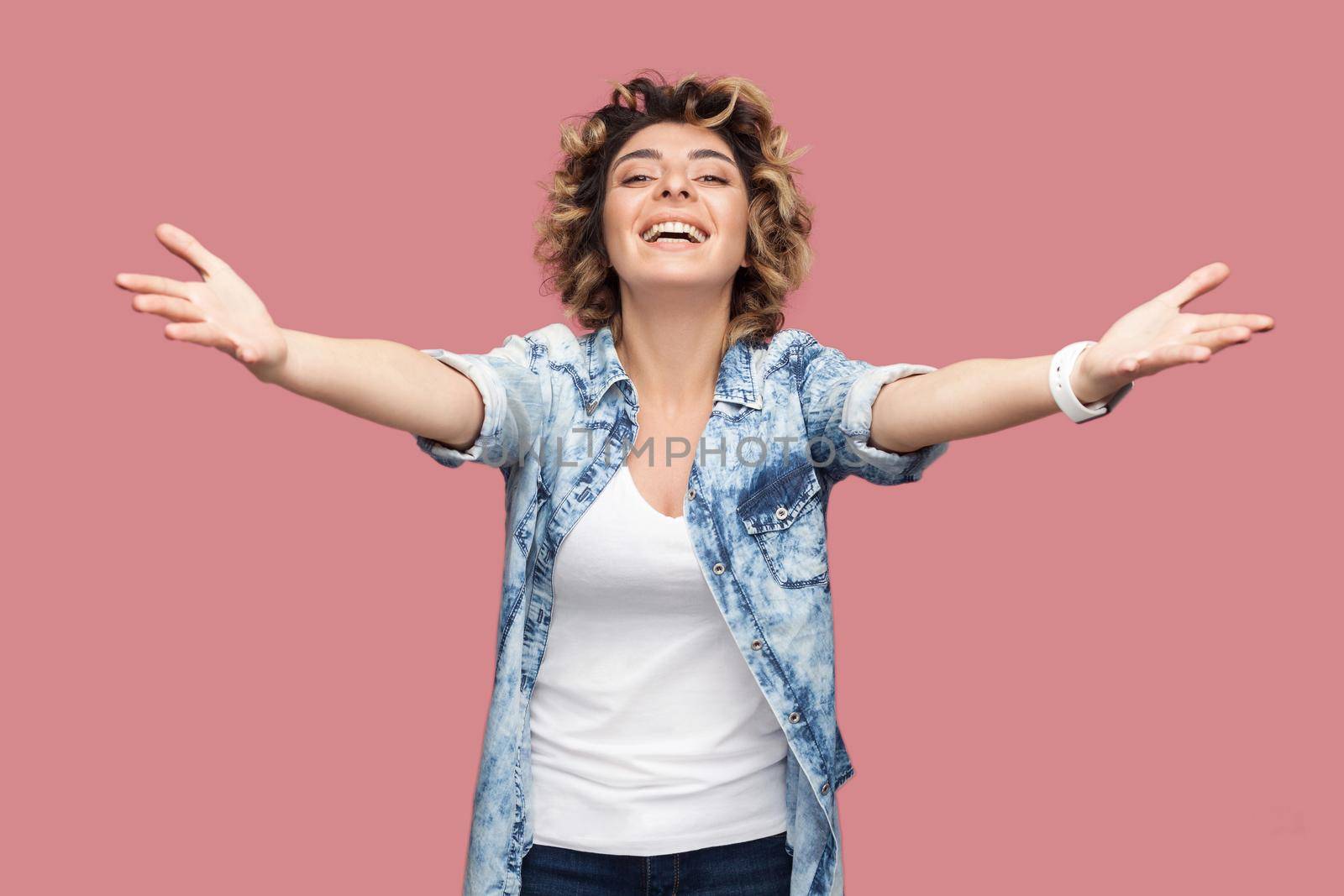 Give me hug. Portrait of happy young woman with curly hair in casual blue shirt standing with raised arms to embrace and looking at camera, toothy smile. indoor studio shot isolated on pink background