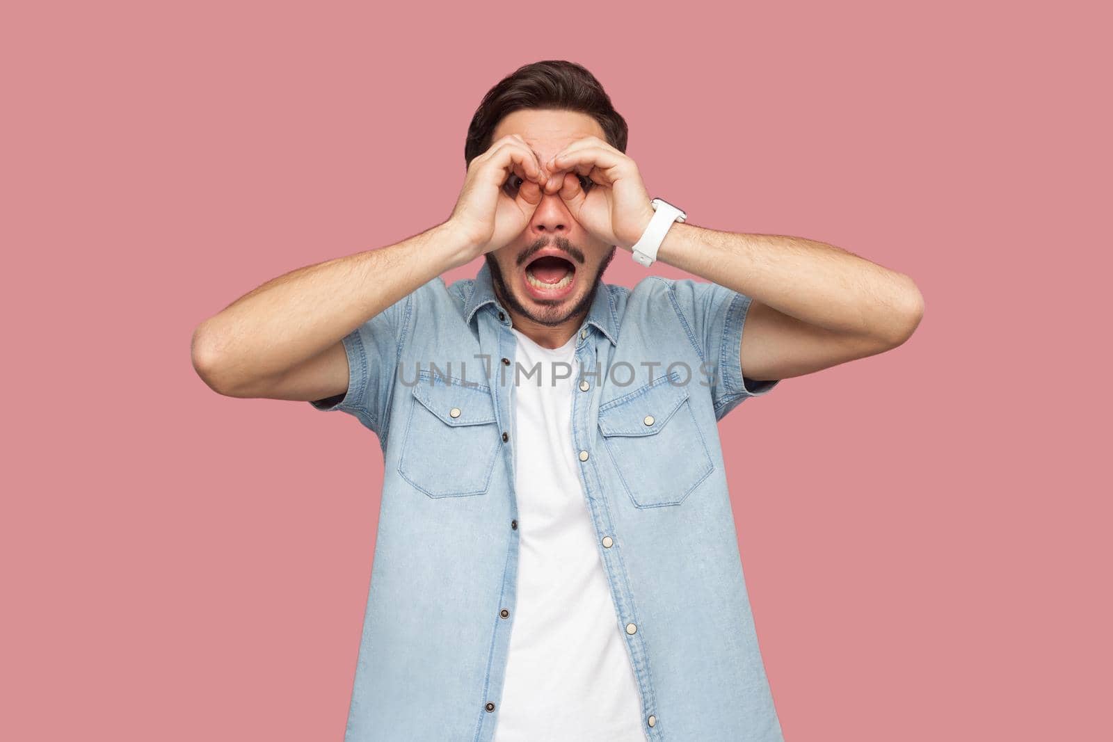 Portrait of surprised or shocked handsome bearded young man in blue casual style shirt standing with binoculars hand gesture on his eyes and looking. indoor studio shot, isolated on pink background.