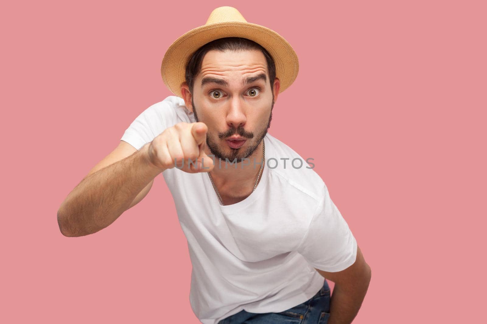 Portrait of shocked bearded young man in white shirt with hat standing, looking and pointing at camera with surprised face. indoor studio shot, isolated on pink background copyspace.