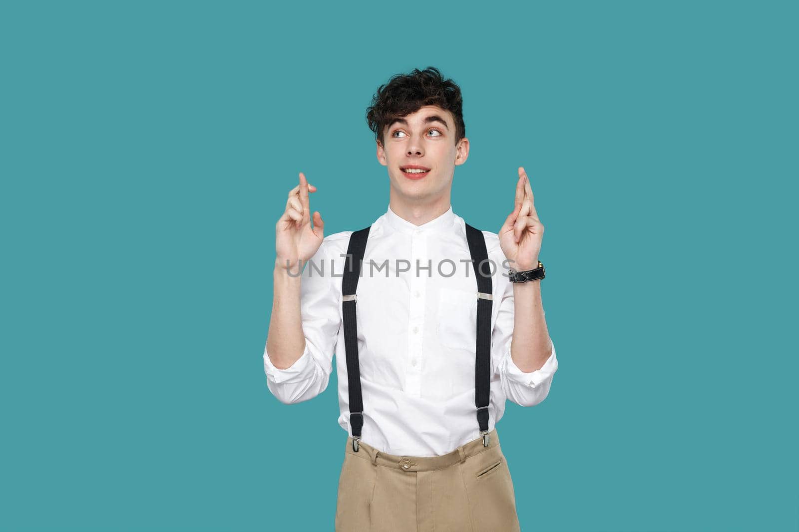 Hopeful man crossed fingers and with to win. Portrait of handsome hipster curly young businessman in classic casual white shirt and suspender standing. indoor studio shot isolated on blue background.