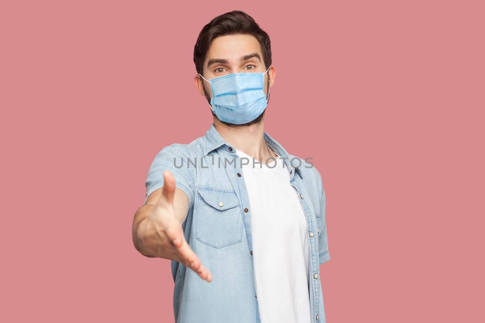 Portrait of happy handsome young man with surgical medical mask in blue shirt standing, looking at camera with smile and giving hand to greeting. indoor studio shot, isolated on pink background.