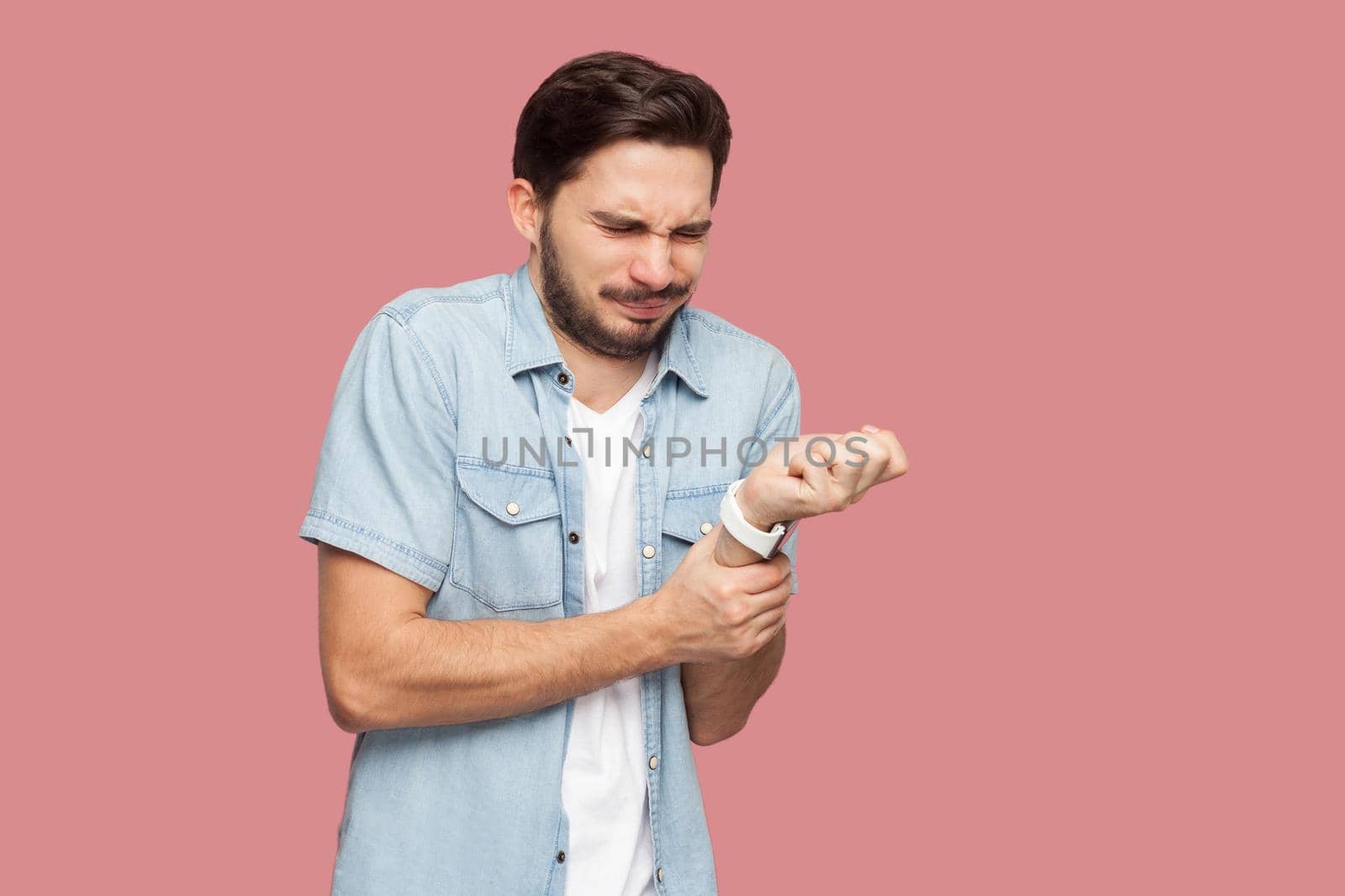 Hand pain. Portrait of sick handsome bearded young man in blue casual style shirt standing and holding his painful hands. indoor studio shot, isolated on pink background.