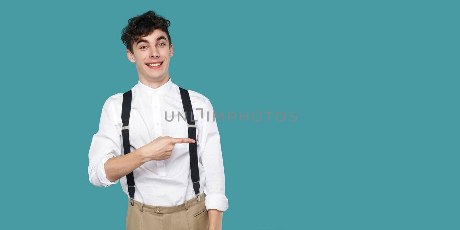 Happy man pointing at empty copyspace and looking at camera with toothy smile. Portrait of hipster curly young businessman in classic casual white shirt suspender. studio shot isolated blue background