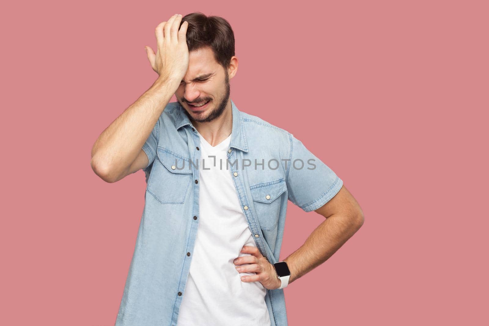 Portrait of sad alone loser bearded young man in blue casual style shirt standing holding his head down and thinking what to do. indoor studio shot, isolated on pink background.