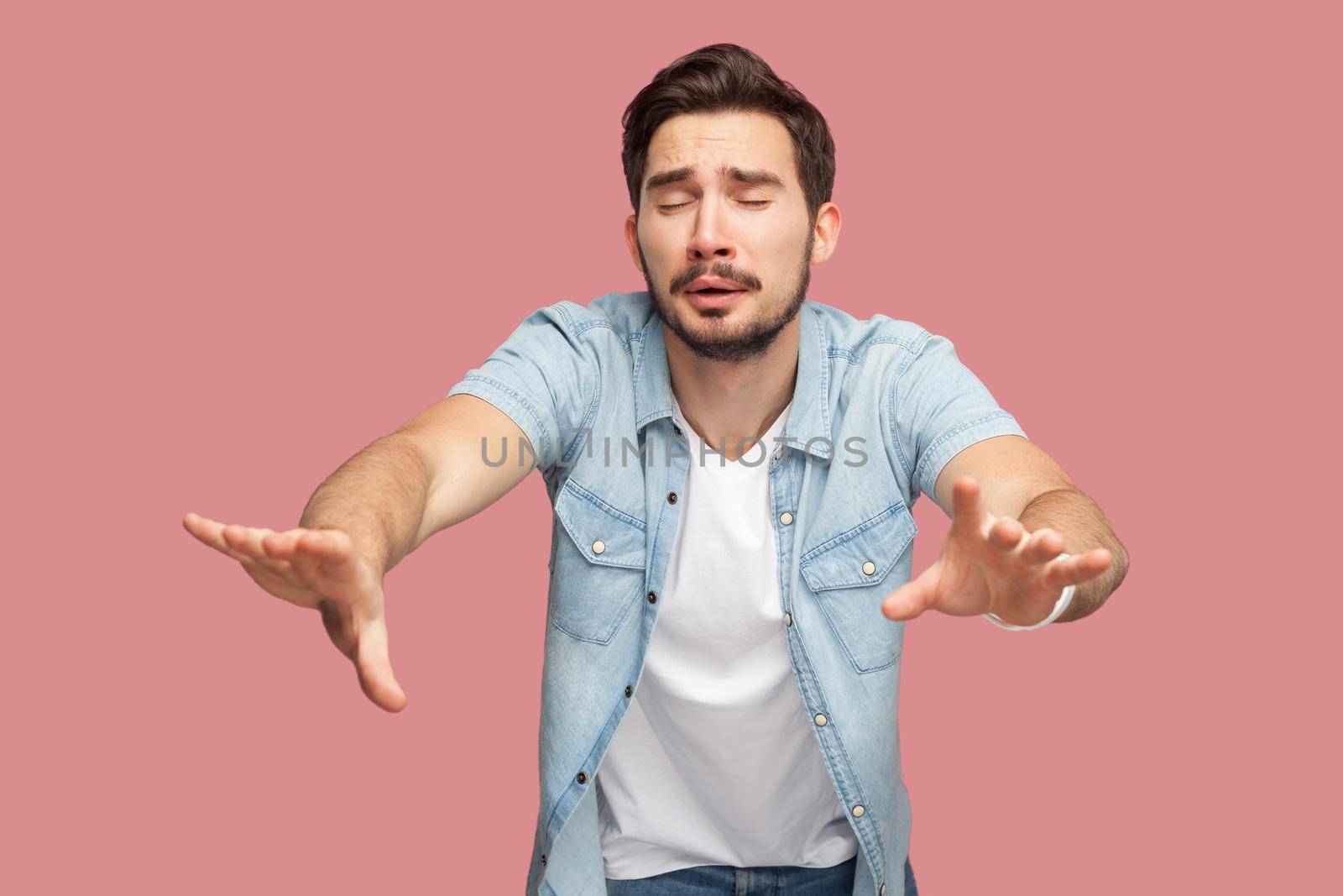 Loneless and blindness. Portrait of lost worry bearded young man in blue casual shirt standing with closed eyes and try to touching something or find. indoor studio shot, isolated on pink background.