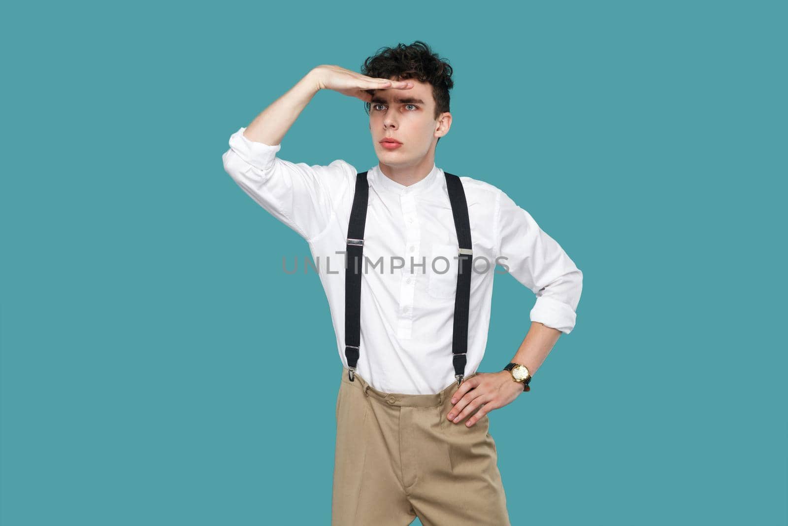 Attentive man looking to far with hand on forrhead. Portrait of handsome hipster curly young businessman in classic casual white shirt and suspender standing. studio shot isolated on blue background.