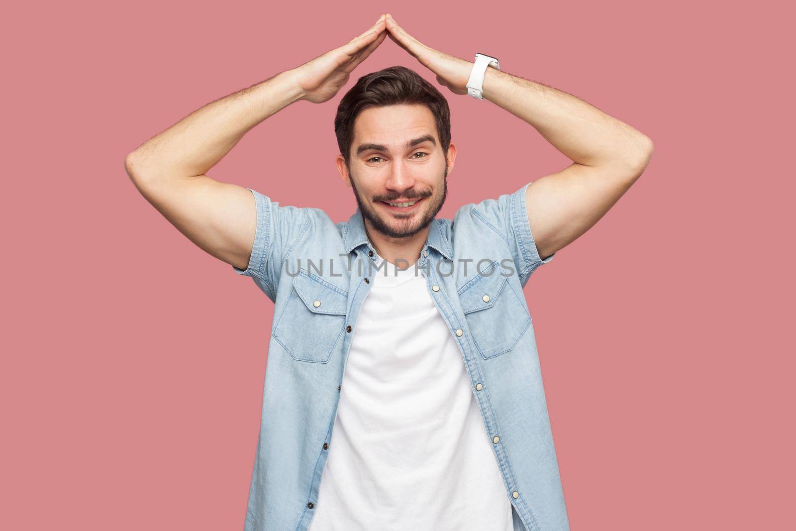 Portrait of happy satisfied handsome bearded young man in blue casual shirt standing with roof gesture hands on head and looking at camera with toothy smile. studio shot, isolated on pink background.