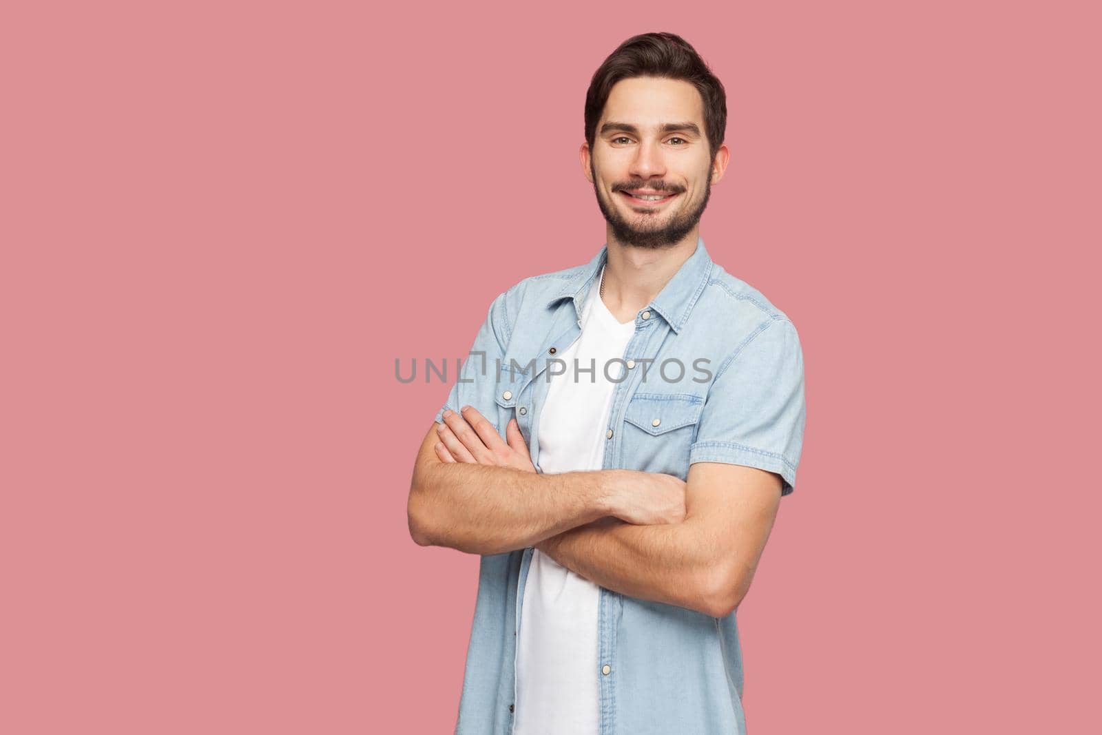 Portrait of happy successful handsome bearded young man in blue casual style shirt standing, raised arms and looking at camera with toothy smile. indoor studio shot, isolated on pink background.