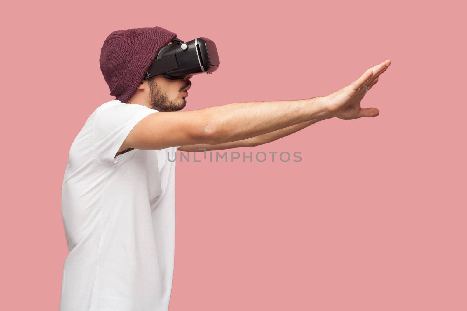 Side view of portrait of wondering bearded young hipster man in white shirt and casual hat standing, wearing vr playing video game with raised arms. Indoor, isolated,studio shot, pink background