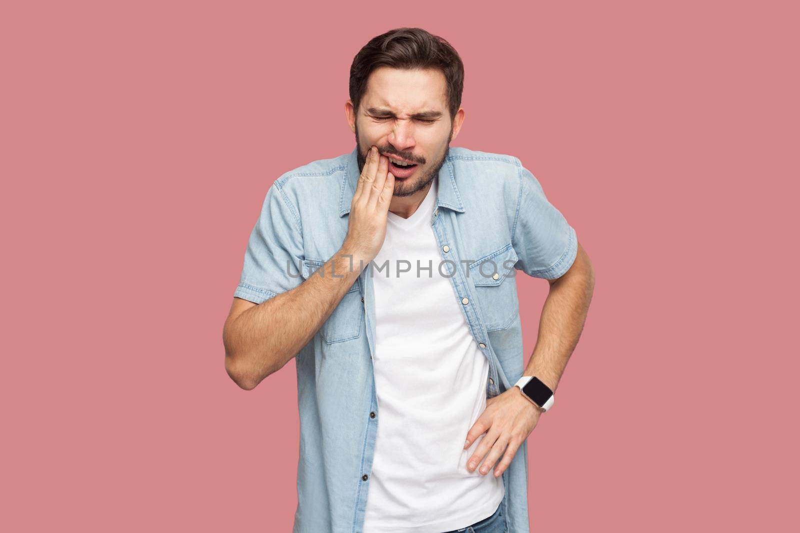 toothache or pain. Portrait of sad sick bearded young man in blue casual style shirt standing and touching his chik because feeling pain on tooth. indoor studio shot, isolated on pink background.