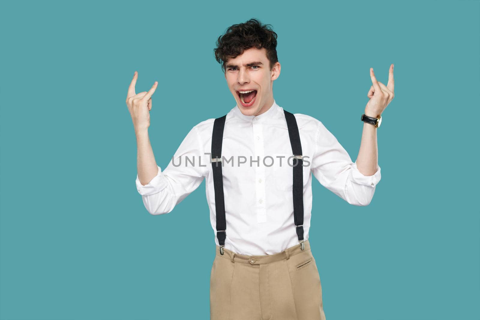 Man screaming and showing rock sign gesture. Portrait of handsome hipster curly young businessman in classic casual white shirt and suspender standing. indoor studio shot isolated on blue background.
