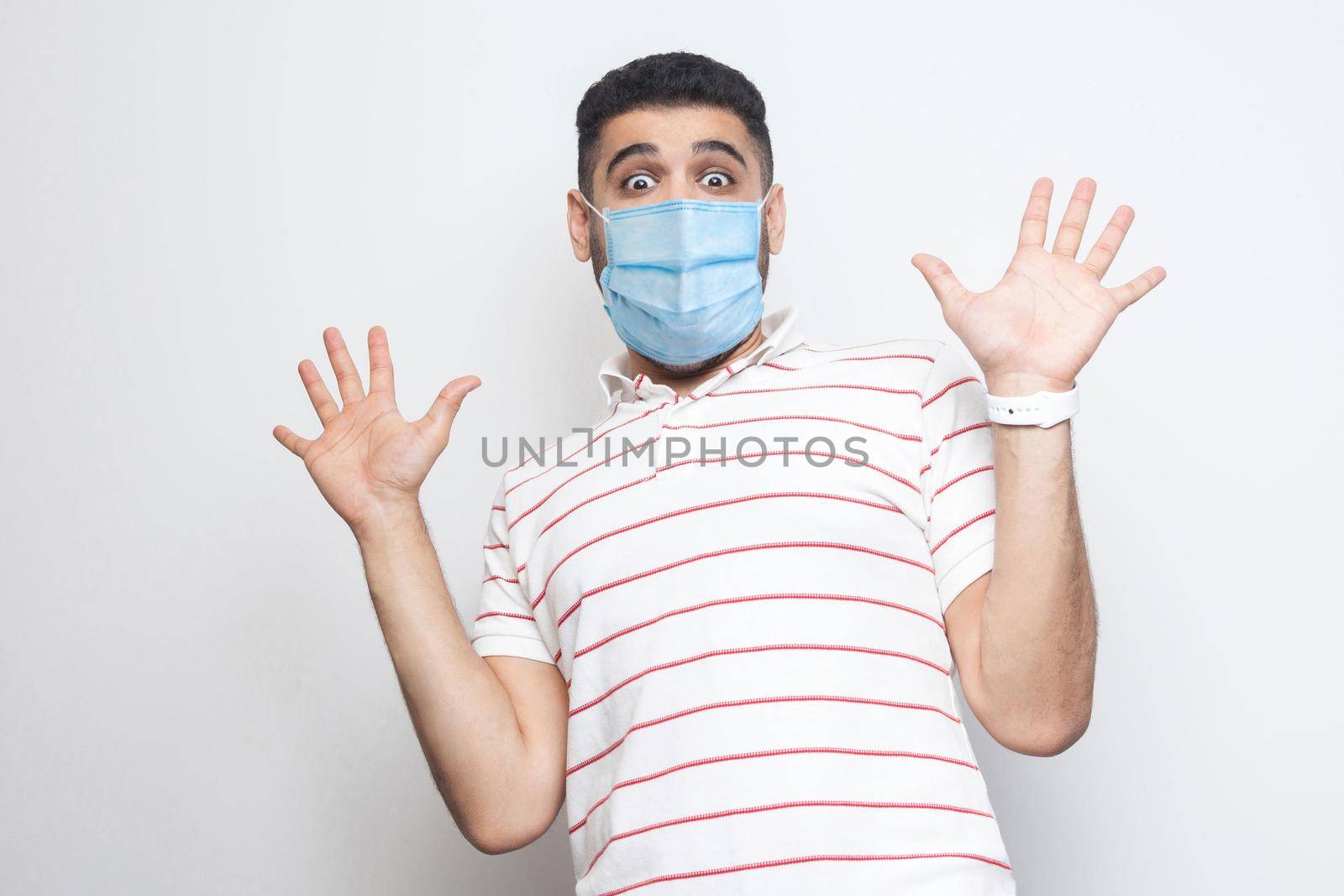 Portrait of shocked young with surgical medical mask man in striped t-shirt standing with raised arms and looking at camera with big eyes and panic. indoor studio shot, isolated on white background.