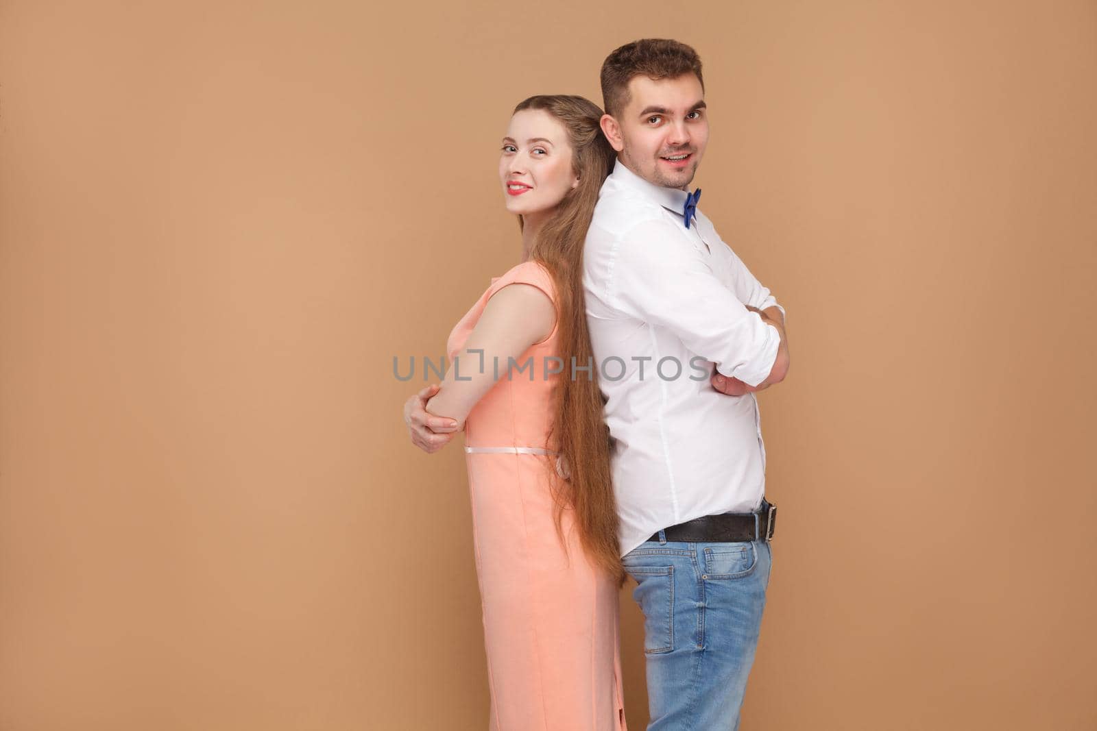 Profile side view of back to back man in casual style and beautiful woman in pink dress, looking at camera with toothy smile and crossed hands. indoor studio shot, isolated on brown background.
