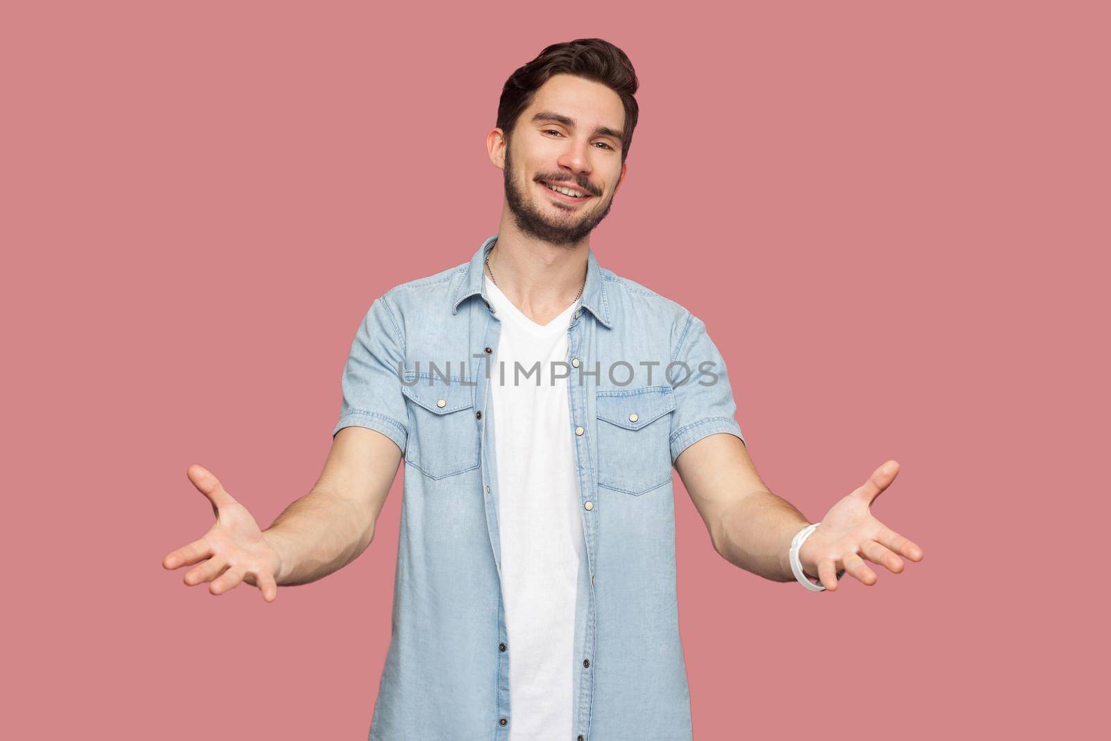Portrait of happy handsome bearded young man in blue casual style shirt standing with raised arms and looking at camera with toothy smile. indoor studio shot, isolated on pink background.