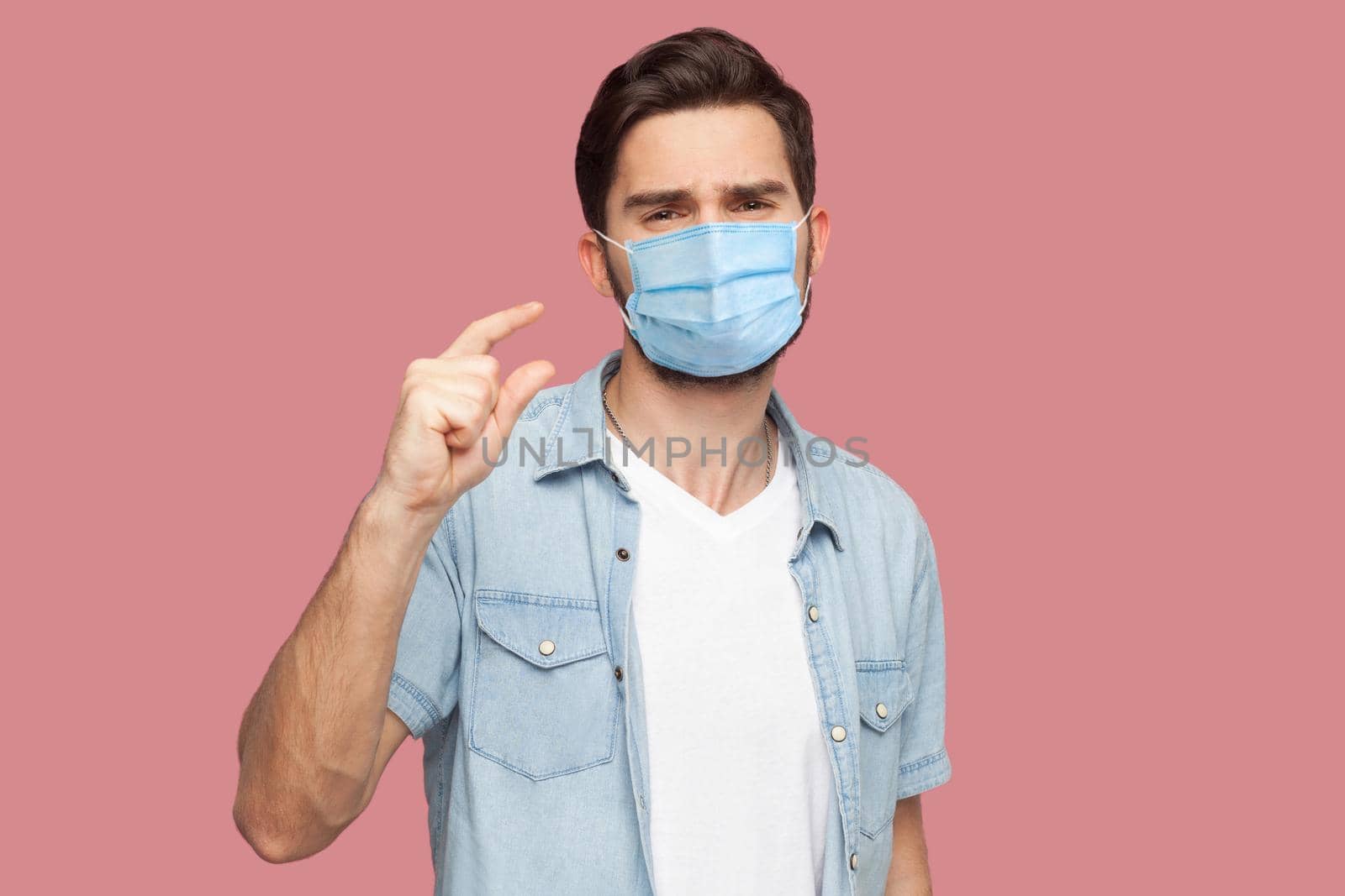 I need a few more. Portrait of young man with surgical medical mask in blue casual style shirt standing and looking at camera and begging for something. indoor studio shot, isolated on pink background