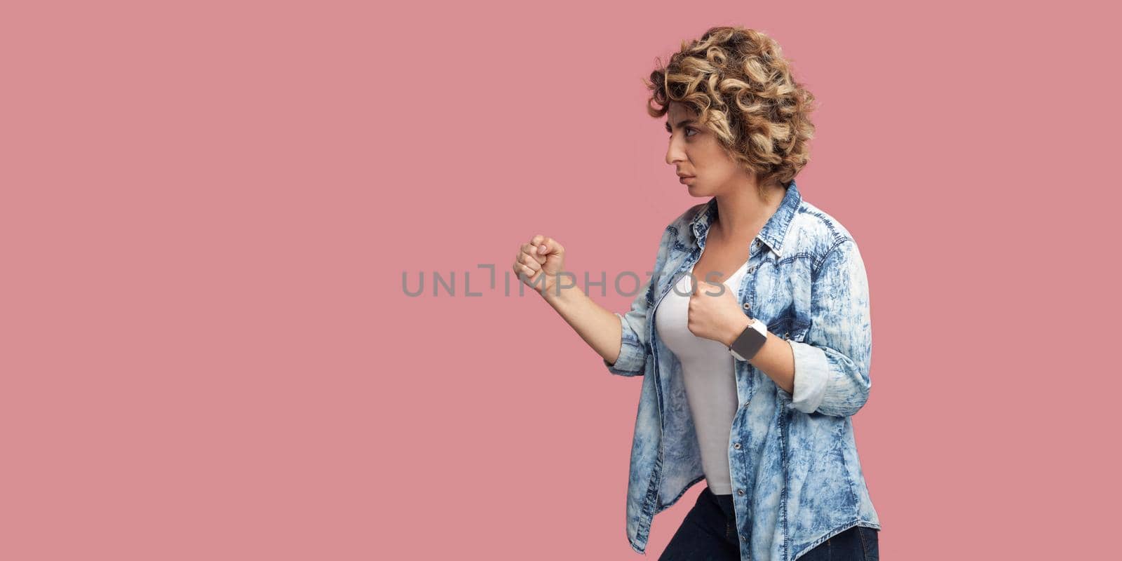 Profile side view portrait of serious young woman with curly hairstyle in casual blue shirt standing with boxing fists, looking forward, ready to attack. indoor studio shot isolated on pink background