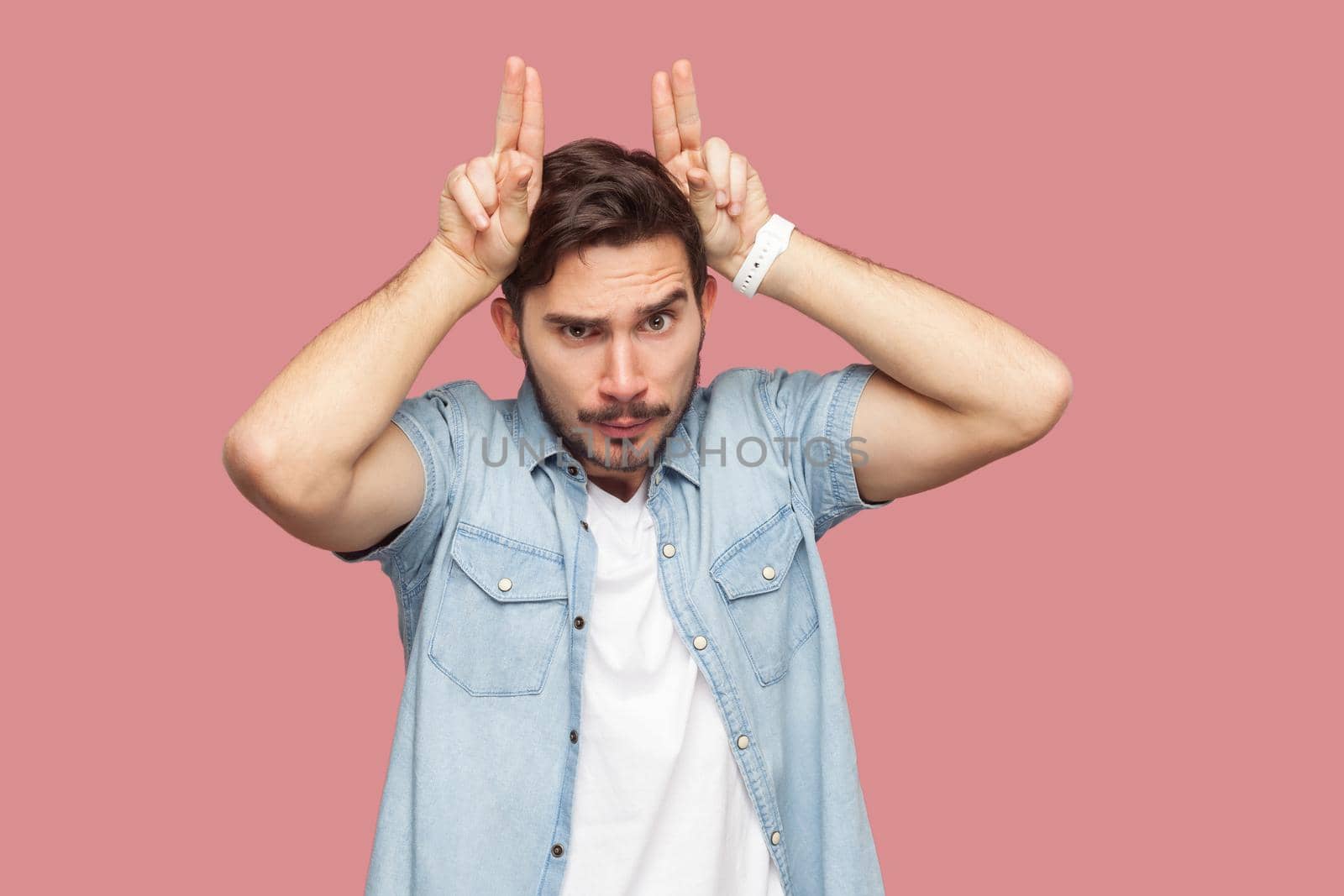 Portrait of angry handsome bearded young man in blue casual shirt standing with horns hand gesture on head and looking at camera with serious face. indoor studio shot, isolated on pink background.