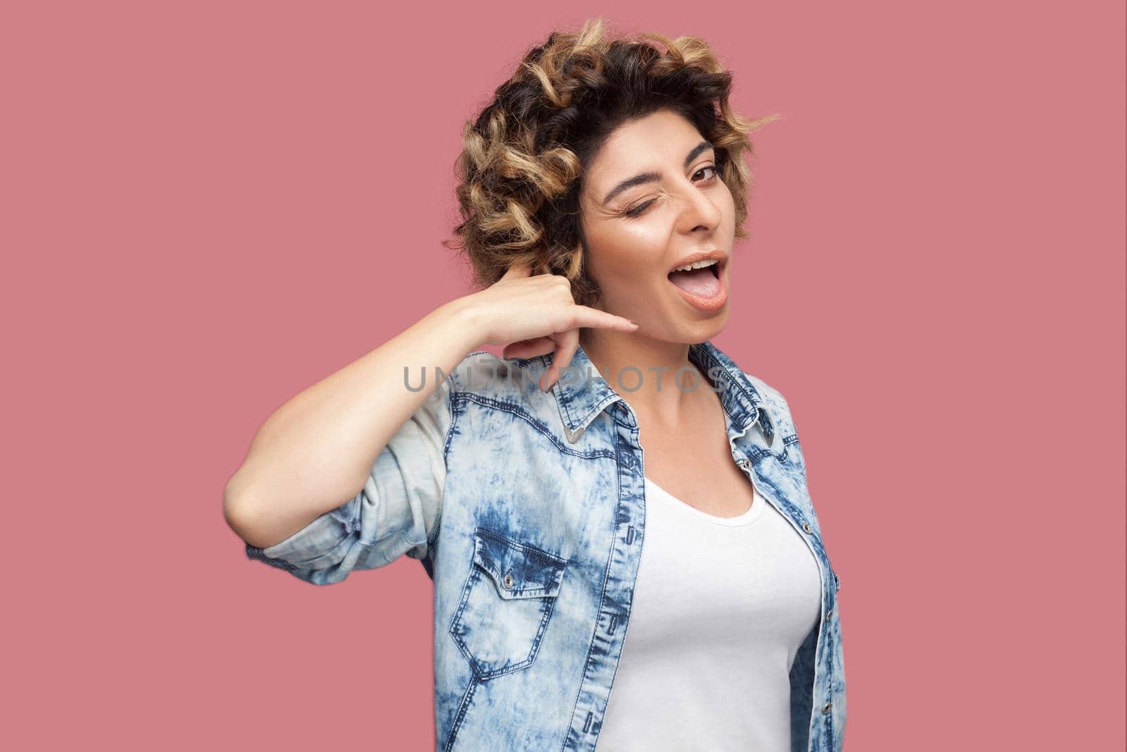 Call me. Portrait of funny young woman with curly hairstyle in casual blue shirt standing with call gesture and looking at camera with funny face. indoor studio shot, isolated on pink background.
