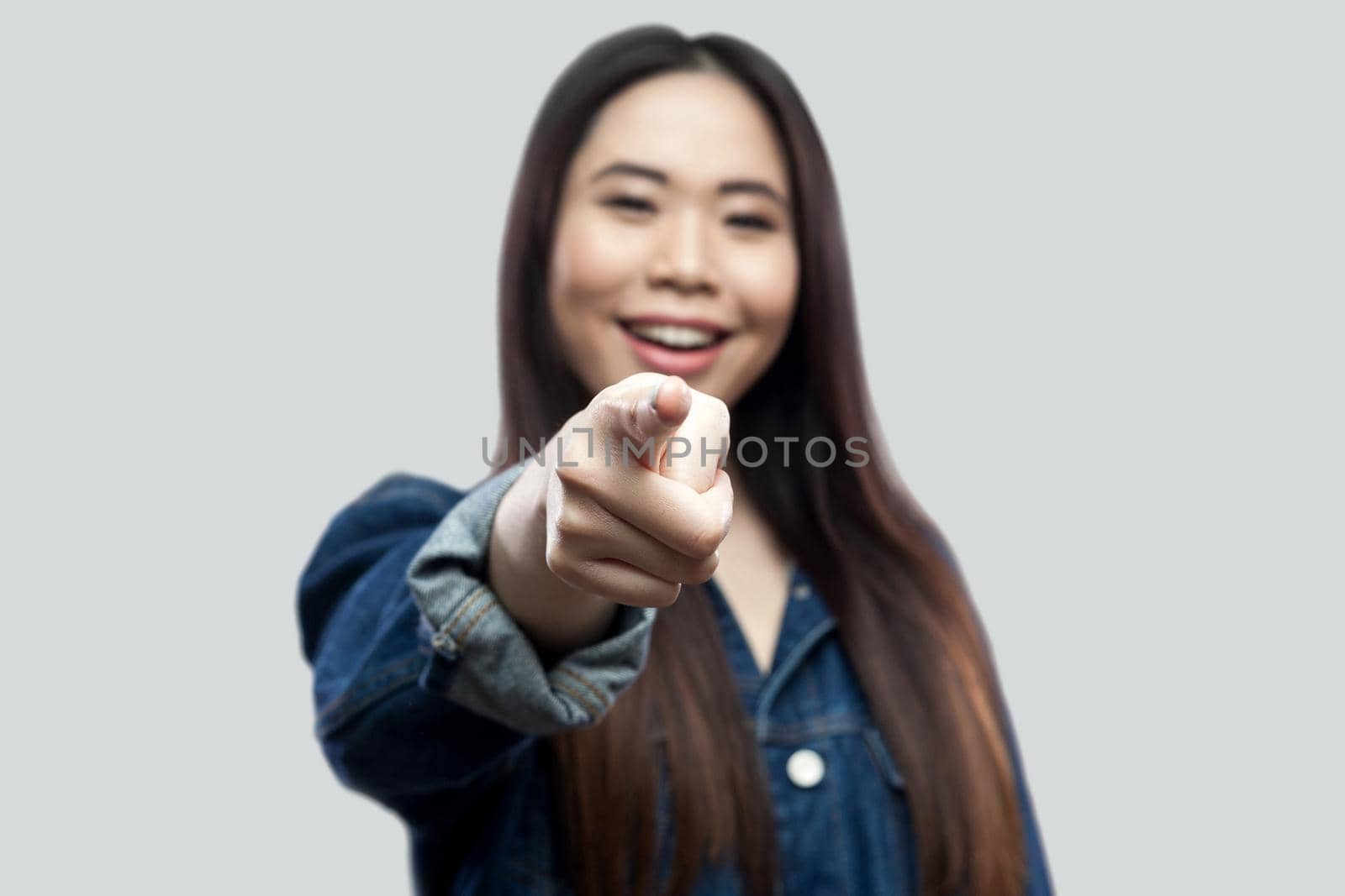 Portrait of excited beautiful brunette asian young woman in casual blue denim jacket with makeup standing toothy smiling and pointing at camera. indoor studio shot, isolated on light grey background.