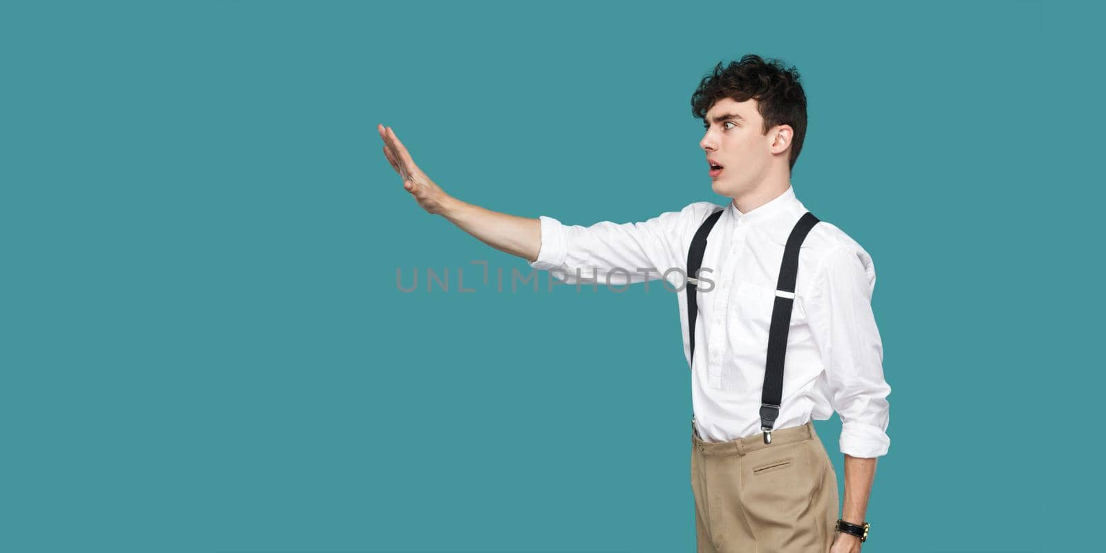 Profile side view portrait of serious handsome hipster curly young businessman in classic casual white shirt and suspender standing with stop sign gesture. studio shot isolated on blue background.